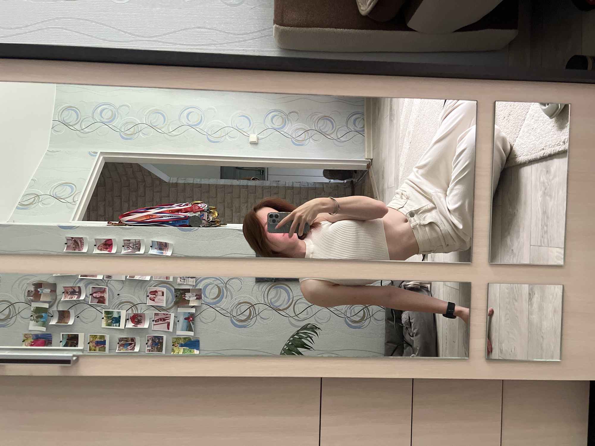 A young woman with reddish-brown hair is taking a selfie in a mirror. She is wearing a white ribbed crop top and off-white pants. She is sitting on the floor with her legs crossed. The mirror is framed by a wooden closet door. The background shows a room with patterned wallpaper, framed photos on the wall, and a doorway leading to another room. There are also medals hanging on a hook on the wall near the doorway. The image appears to be taken indoors, likely in a residential setting in Chaikovsky, Russia.