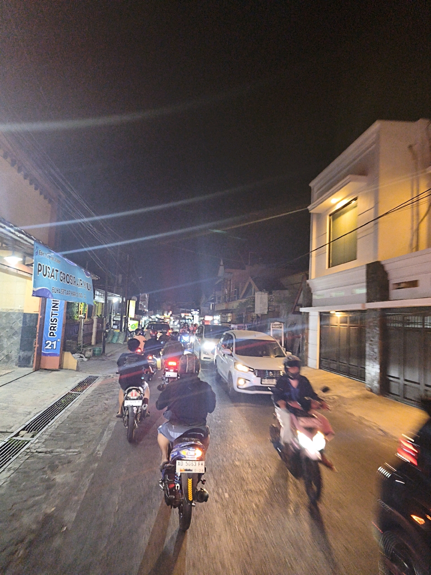 In Cemani, Indonesia, this image captures a nighttime scene on a street filled with traffic. The perspective is from behind a group of motorcyclists. Dominating the left side of the frame is a blue banner with Indonesian text, including "PUSAT GROSIR AIR MINUM" (Water Wholesale Center) and "BUKA SETIAP HARI 10.00-17.00" (Open Every Day 10 AM - 5 PM). Below this, a vertical banner displays "PRISTINI" and "21".
The street is paved and has drainage grates on the left. The road is congested with various vehicles, primarily motorcycles, with their headlights and taillights illuminating the scene. A white car is visible in the mid-ground, and the building on the right is a modern, light-colored structure with illuminated windows and a large dark gate. The overall atmosphere is that of a busy commercial street at night.