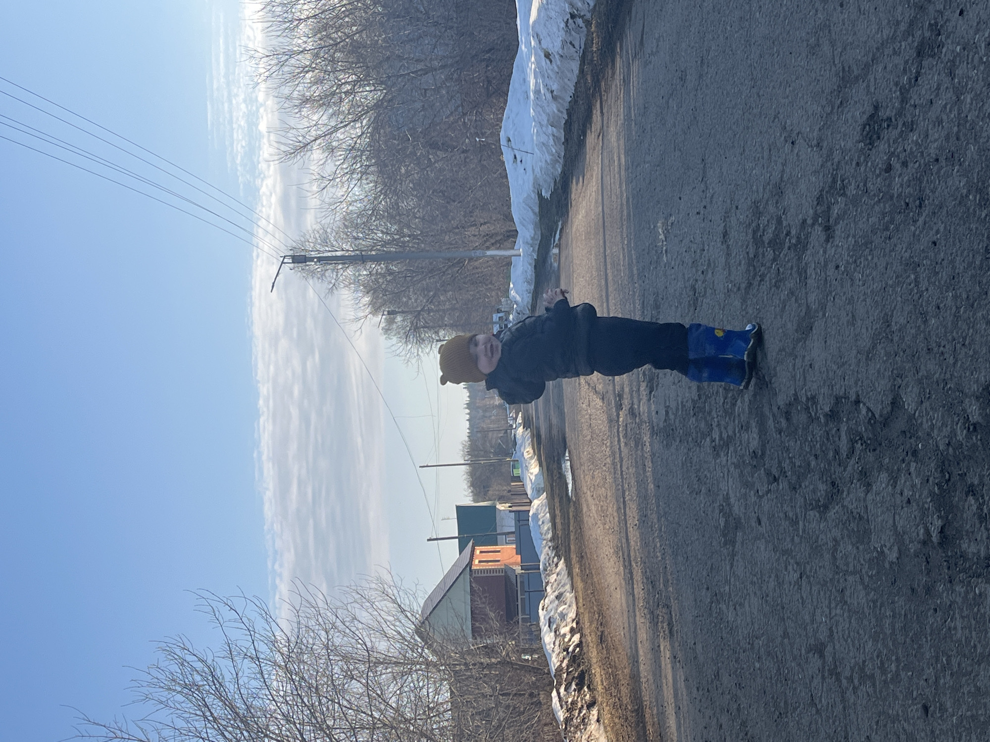 A young child stands on a paved road in the city of Павловка, Russia. The child wears a dark jacket, dark pants, a brown hat, and blue boots. Patches of snow line the side of the road. In the background, there are trees without leaves, utility poles with wires, and some buildings. The sky is blue with streaky clouds.