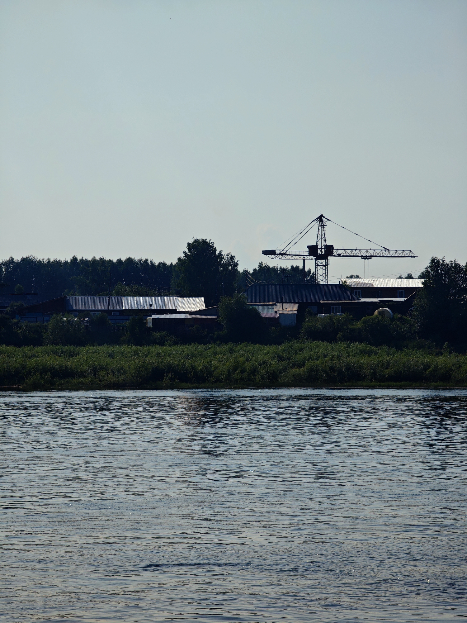 This is a wide shot of a body of water with a village and a construction crane on the opposite bank, located in the village of Verkhne-Pashino, Russia. The water is calm with gentle ripples, reflecting the sky and the bank. The bank is lined with dense green foliage and trees. Behind the foliage, several buildings are visible, including what appears to be a long, corrugated metal-roofed structure and other houses. A large construction crane stands prominently behind the buildings, its silhouette stark against the clear, pale blue sky. The overall scene suggests a rural or semi-industrial area situated near a significant body of water. No people are visible in the image.