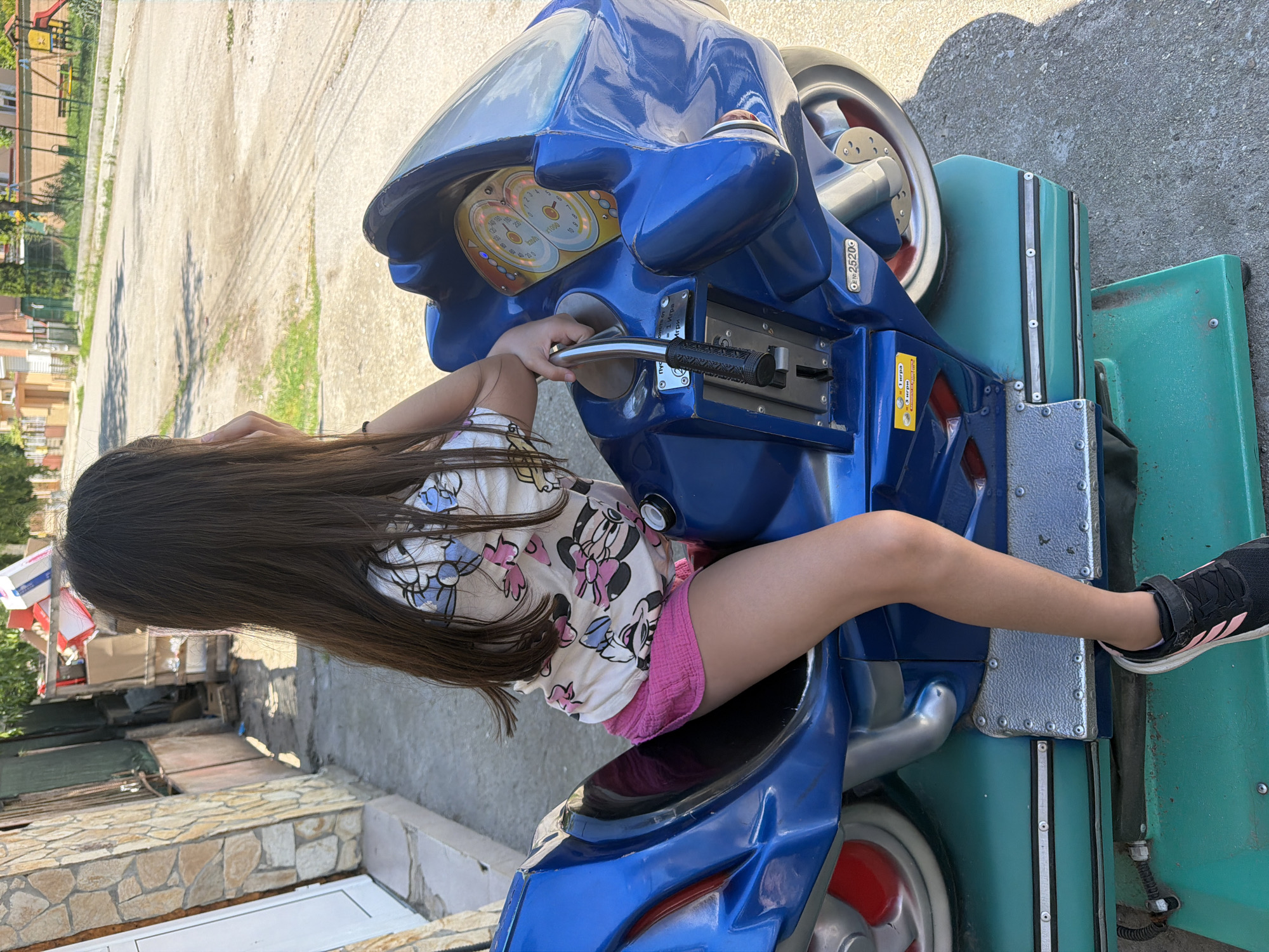 A young girl with long, dark hair sits on a blue kiddie ride shaped like a motorcycle. She is wearing a white t-shirt with a Minnie Mouse print and pink shorts. Her right hand rests on the handlebars, and her left hand is near her face. The motorcycle ride is situated outdoors on a paved surface. In the background, there is a building with a stone facade, a fence, and a street with houses and trees. The setting appears to be in the city of Ravno pole, Bulgaria, as indicated by the text on the ride which includes Cyrillic script.