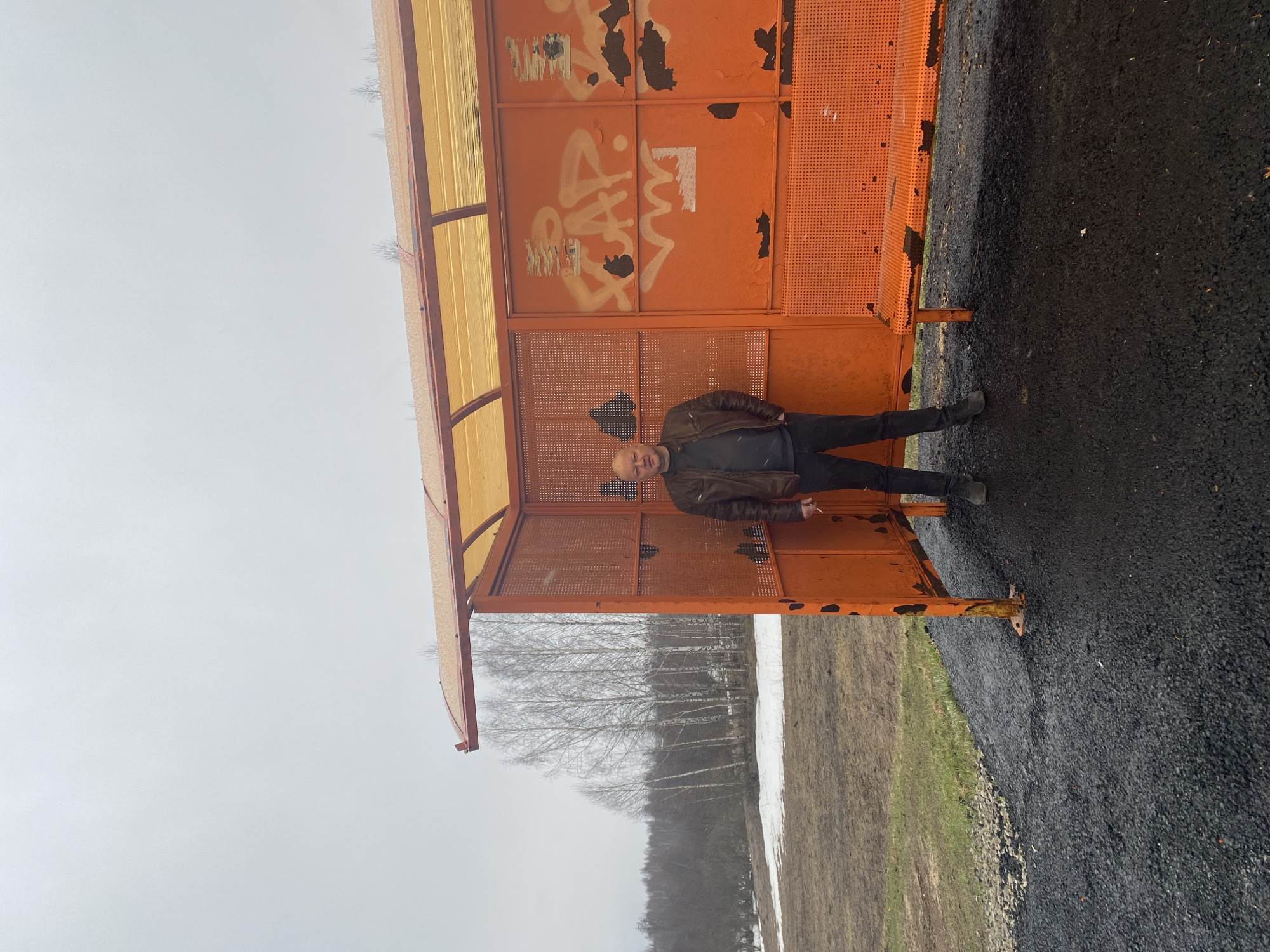 A man stands within an orange bus shelter in what appears to be Буль-Кайпаново, Russia. The shelter features graffiti and some rust. The man wears a brown jacket and dark trousers. Outside the shelter, the ground is paved on one side and transitioning to a field with snow and bare trees on the other. The sky is overcast.