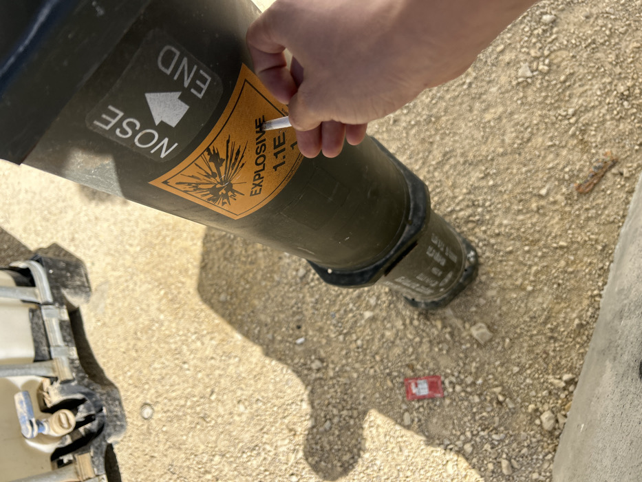 A person holds a cigarette to an explosive ordnance. The device is olive green with a yellow label marked "EXPLOSIVE 1.1E," and an arrow indicating the "NOSE END." The device sits on dirt with small rocks. A red object is near the base of the device, with a section of a white container and its metal frame.