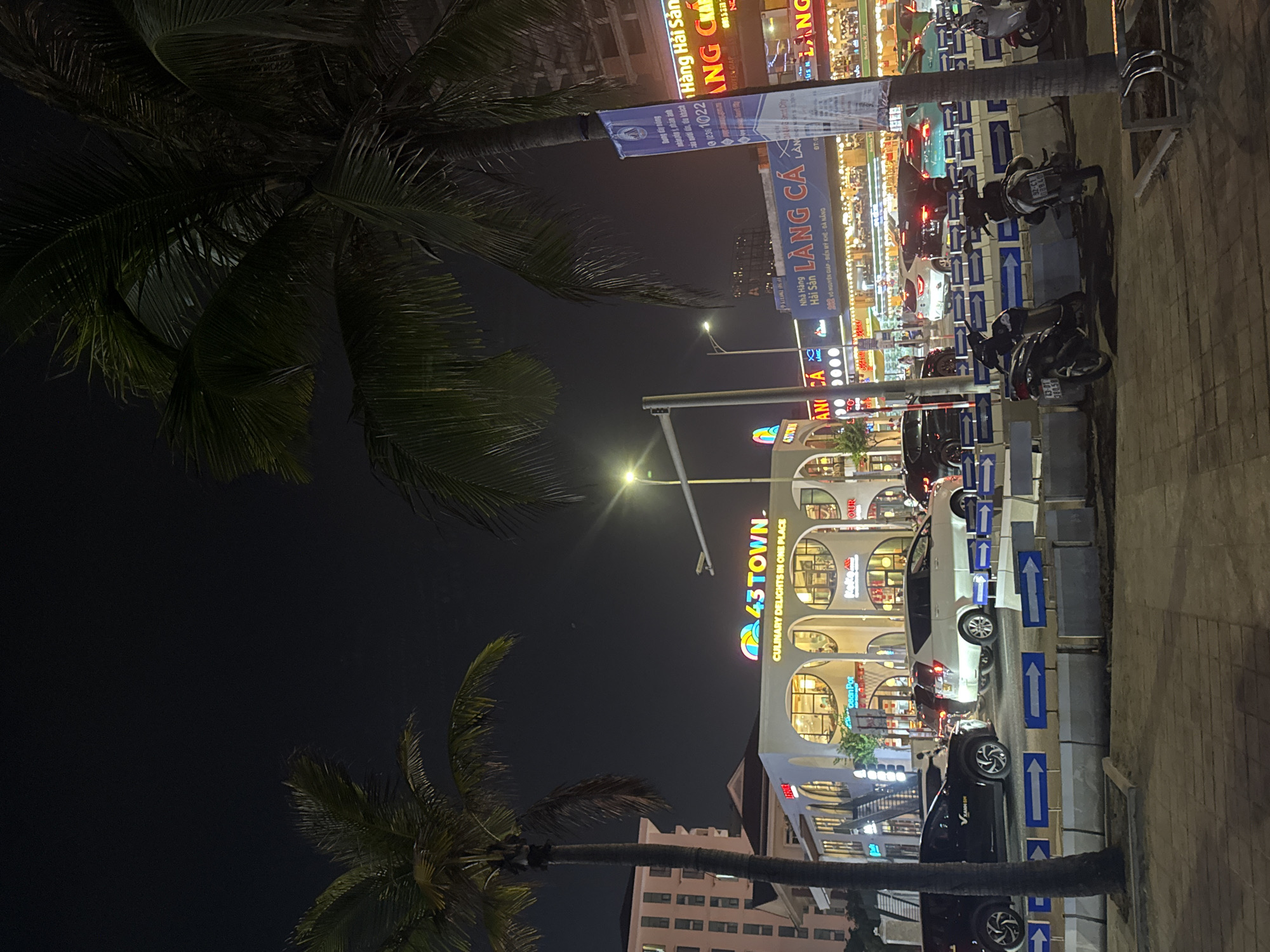 This outdoor night scene captures a street in Đà Nẵng, Vietnam. A palm tree extends into the left side of the frame, contrasting with the illuminated structures on the right. A tall building with multiple levels of arched windows and signs reading "43 TOWN. CULINARY DELIGHTS IN ONE PLACE" and "Ocean Pot" stands prominently. Other signs in Vietnamese are visible higher up, including "Hàng Hải Sản" and "LÀNG CÁ." Parked cars and motorbikes line the street, separated by blue and white arrow markers. Streetlights illuminate the scene. The overall composition provides a glimpse into the urban nightlife of Đà Nẵng.