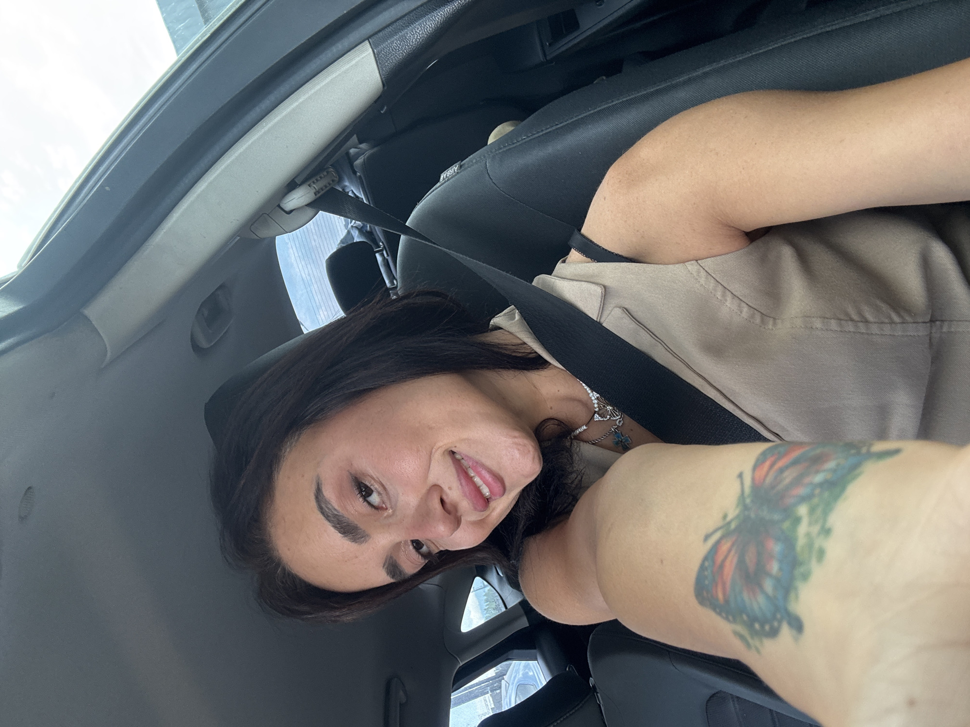 A woman with dark hair and fair skin, wearing a beige vest and a necklace, is seen from the chest up, taking a selfie inside a car. She is smiling and looking at the camera. Her left arm is extended towards the camera, revealing a colorful butterfly tattoo. The interior of the car, including the seatbelt and the driver's seat, is visible. The background shows a glimpse of the sky and possibly other vehicles. The image appears to have been taken during the day. The scene is set in the city of Tbilisi, Georgia.