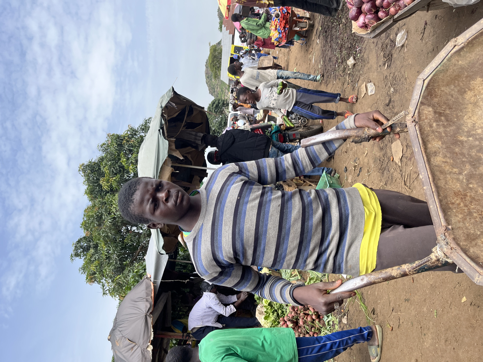 A young Black male, likely a vendor, stands in a busy outdoor market. He is wearing a grey, blue, and black striped long-sleeved shirt with a bright yellow undershirt peeking out. He is holding a metal handle, possibly part of a wheelbarrow or cart, with his right hand. His expression is neutral.
The setting appears to be a bustling marketplace in Gura Top, Nigeria, characterized by dirt ground and makeshift stalls covered with tarps. In the background, other people are visible, some appearing to be shoppers or other vendors. Several stalls are laden with produce, including piles of red onions and leafy green vegetables. A motorcycle is parked nearby, suggesting transportation. The sky is blue with scattered white clouds, indicating daytime with fair weather. The overall scene depicts the lively atmosphere of a local market. There is no visible text in the image.