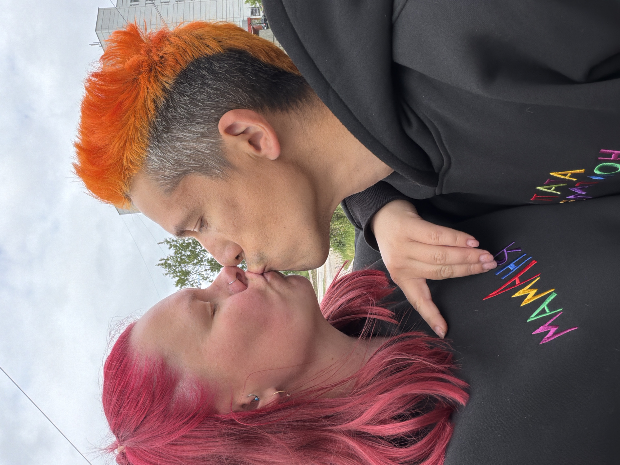 An outdoor image depicts two individuals engaged in a kiss under an overcast sky in the city of Chaikovsky, Russia. The person on the left has bright pink hair and wears a silver nose ring and a blue stud in their ear. They are dressed in a black hoodie featuring multicolored embroidered Cyrillic text spelling "МАМАНИЯ" (MAMANIYA). Their eyes are closed, and their right hand rests on the left arm of the other individual. The person on the right features vibrant orange hair styled with shaved gray sides. They are also wearing a black hoodie, with visible multicolored embroidered Cyrillic letters forming "ПАТА" and "M'OH," part of the word "ПАТАМОН" (PATAMON). Their eyes are closed as they kiss the individual on the left. The background consists of a light gray, cloudy sky, traversed by a thin utility wire. To the left, green tree foliage is visible, while a multi-story building with numerous windows occupies the right side of the background, indicating an urban setting.
