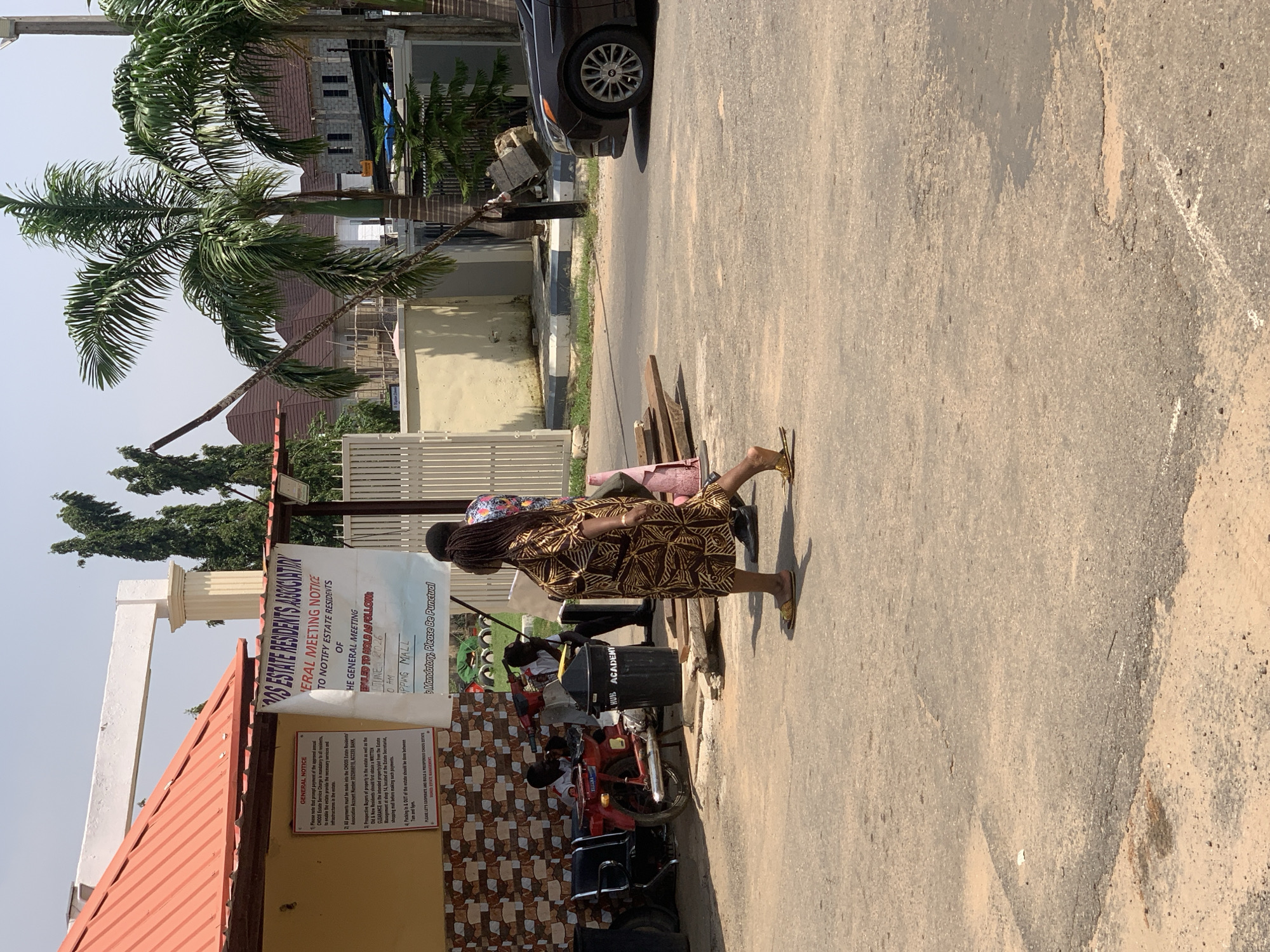 The image shows a street scene in Abuja, Nigeria. In the foreground, a woman wearing a patterned dress and yellow sandals walks away from the camera. Behind her, a black sedan is parked on the right side of the road, with its front end facing towards the viewer. Further down the street, a red motorcycle is parked, with two individuals sitting on it. A black trash bin labeled "HUB ACADEMY" is positioned next to the motorcycle.
On the left side of the image, a building with a red roof and yellow walls is visible. A sign is posted on the wall, containing text related to "GENERAL NOTICE" and "CHIDOE ESTATE SERVICE CHARGE". To the right of this building, a white gate stands open. In the background, palm trees and residential buildings are present, suggesting a suburban or residential area. The overall environment appears to be outdoors on a sunny day, with a paved road surface that shows some signs of wear and tear.