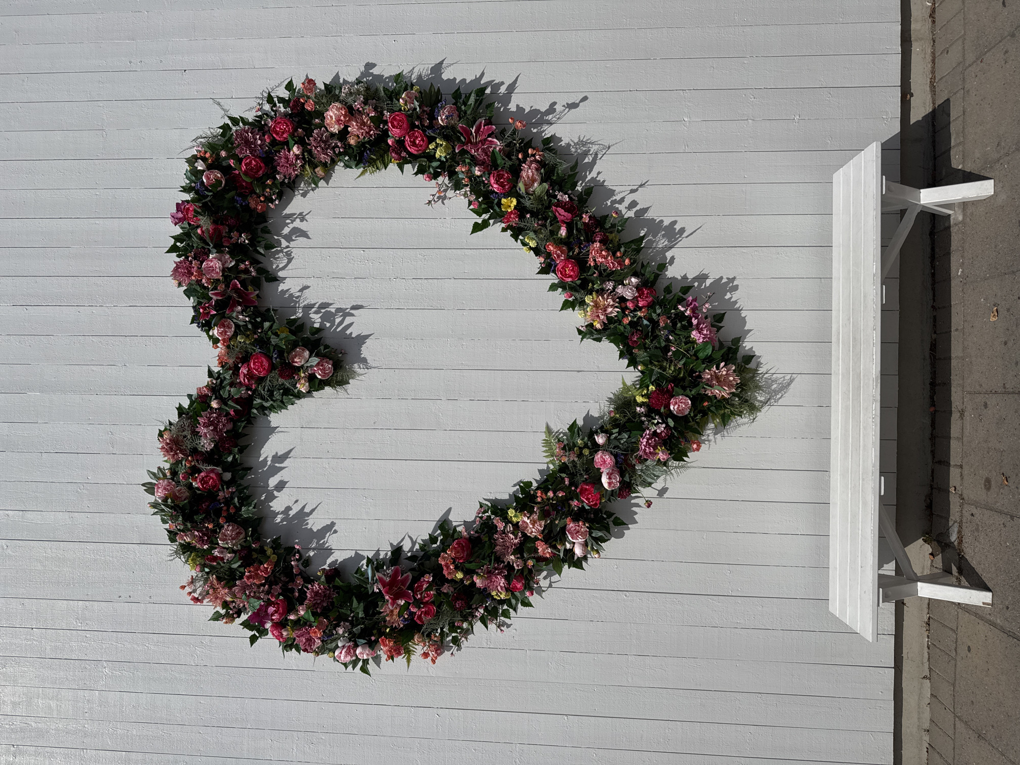 This is a photograph of a large, heart-shaped floral arrangement mounted on a white, slatted wooden wall. The floral arrangement is dense with a variety of flowers in shades of pink, red, and deep magenta, interspersed with dark green foliage. Below the floral heart, a simple white wooden bench with four legs is positioned in front of the wall, casting a shadow on the grey, paved ground. The scene is outdoors, under bright sunlight, as indicated by the sharp shadows. The image appears to be taken at a public space, possibly a park or a street in Järfälla kommun, Sweden, given the context provided.