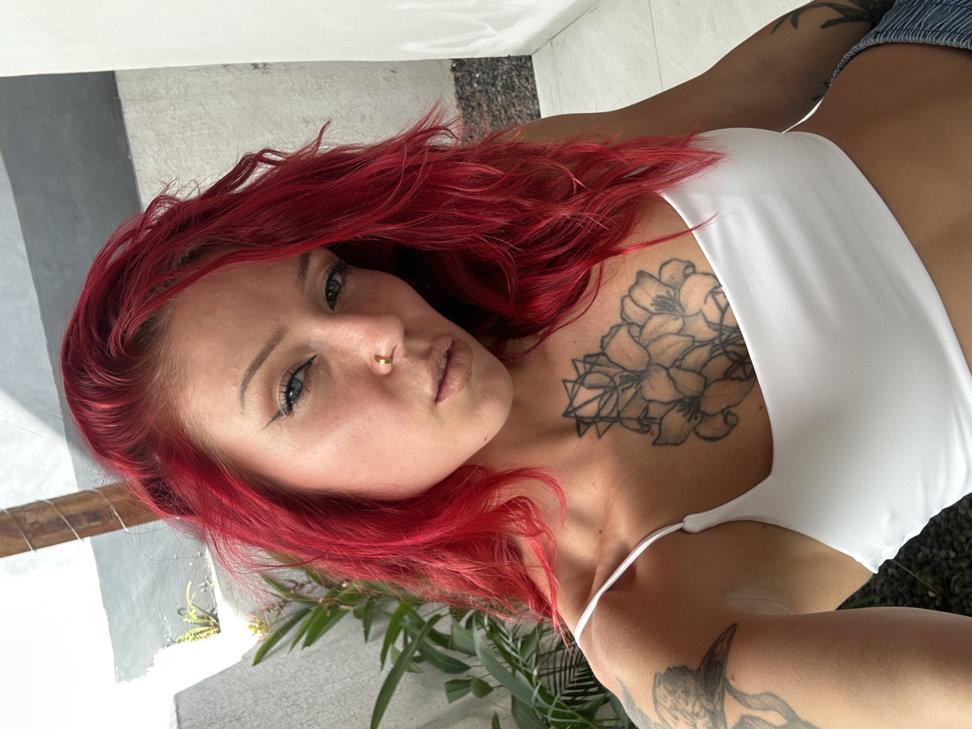 A woman with red hair, a white top, and tattoos is lying down. She has a nose ring and is looking at the camera. The background is a mix of white walls, green plants, and a textured dark surface. The photo was taken in North Kuta, Indonesia.