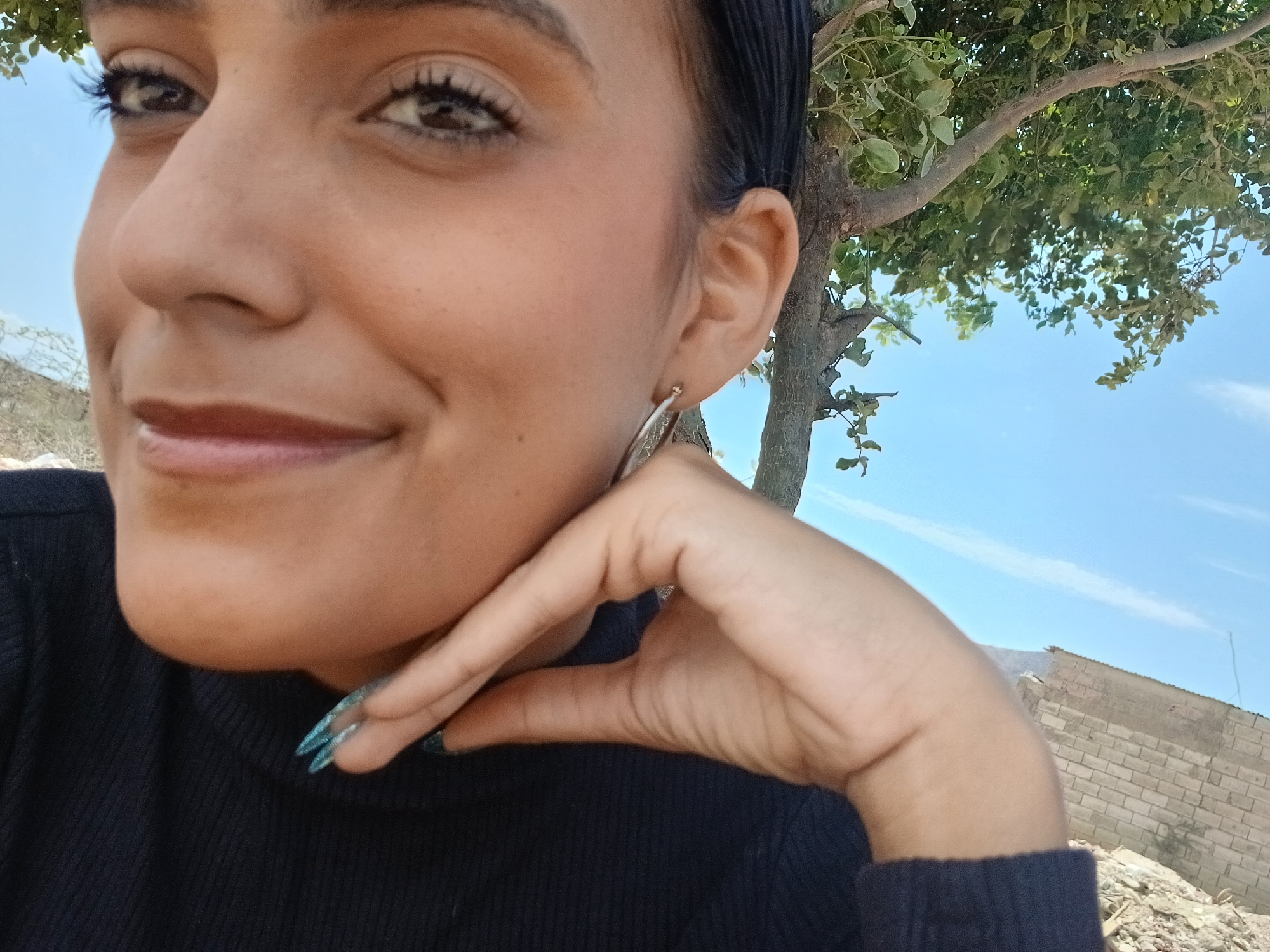 A close-up portrait features a young woman smiling subtly, with her head slightly tilted and her gaze directed towards the viewer. Her dark hair is pulled back, and she has visible eye makeup, including mascara. She is wearing a black, ribbed, long-sleeved top. Her right hand rests gently under her chin, showcasing long, pointed fingernails painted with an iridescent blue-green polish. A silver-colored hoop earring is visible in her right ear.
The setting is outdoors during a bright daytime. Behind and to the right of the woman, a tree with green foliage and branches is prominent. The upper and middle background is dominated by a clear, light blue sky with faint white clouds. Towards the lower right, a low wall made of light-colored concrete blocks is visible, with some debris or rubble at its base, suggesting a rustic or semi-urban environment in Altagracia, Venezuela. The overall mood of the image is calm and pleasant. No text is visible.