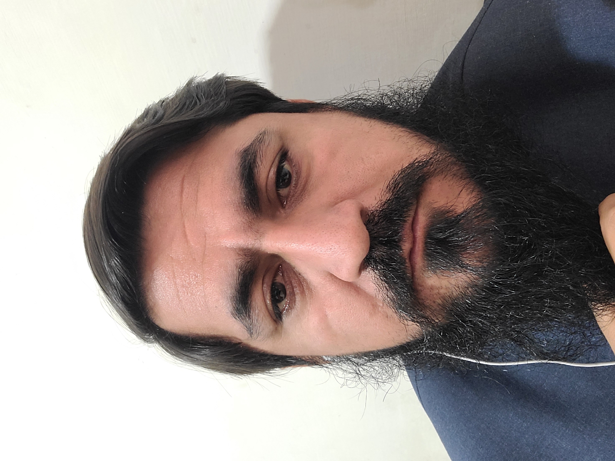 A close-up portrait of a man with dark hair and a full black beard is presented against a plain, light-colored wall. He is wearing a dark blue shirt. A white earbud cable is visible along his neck. The photo appears to be taken indoors. There is no information available to ascertain the location in Quetta City Tehsil, Pakistan.