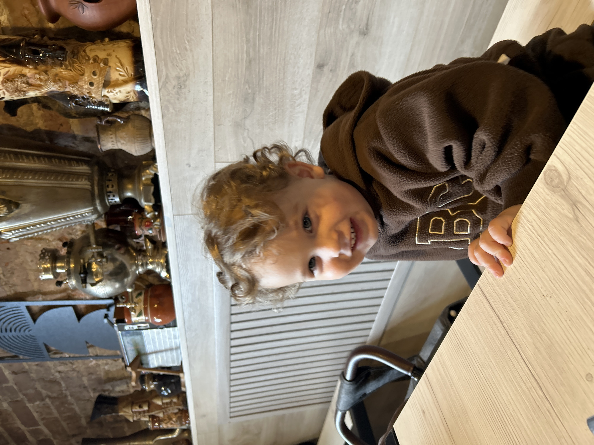 A young child with light brown curly hair and blue eyes is seated at a light-colored wooden table, looking forward and smiling directly at the viewer. The child wears a dark brown fleece hoodie featuring a light-colored "B" or "BP" logo on the chest. To the child's left, a dark-colored stroller or child seat handle is partially visible. The background features a rustic brick wall above a white shelf. The shelf displays a collection of traditional decorative items, including at least two metallic samovars, various ceramic pitchers and vases, and a dark geometric metal wall hanging. Below the shelf and behind the child, a light-colored wooden panel with vertical slats is visible. The specific location within Klin, Russia, cannot be determined from the visual evidence.