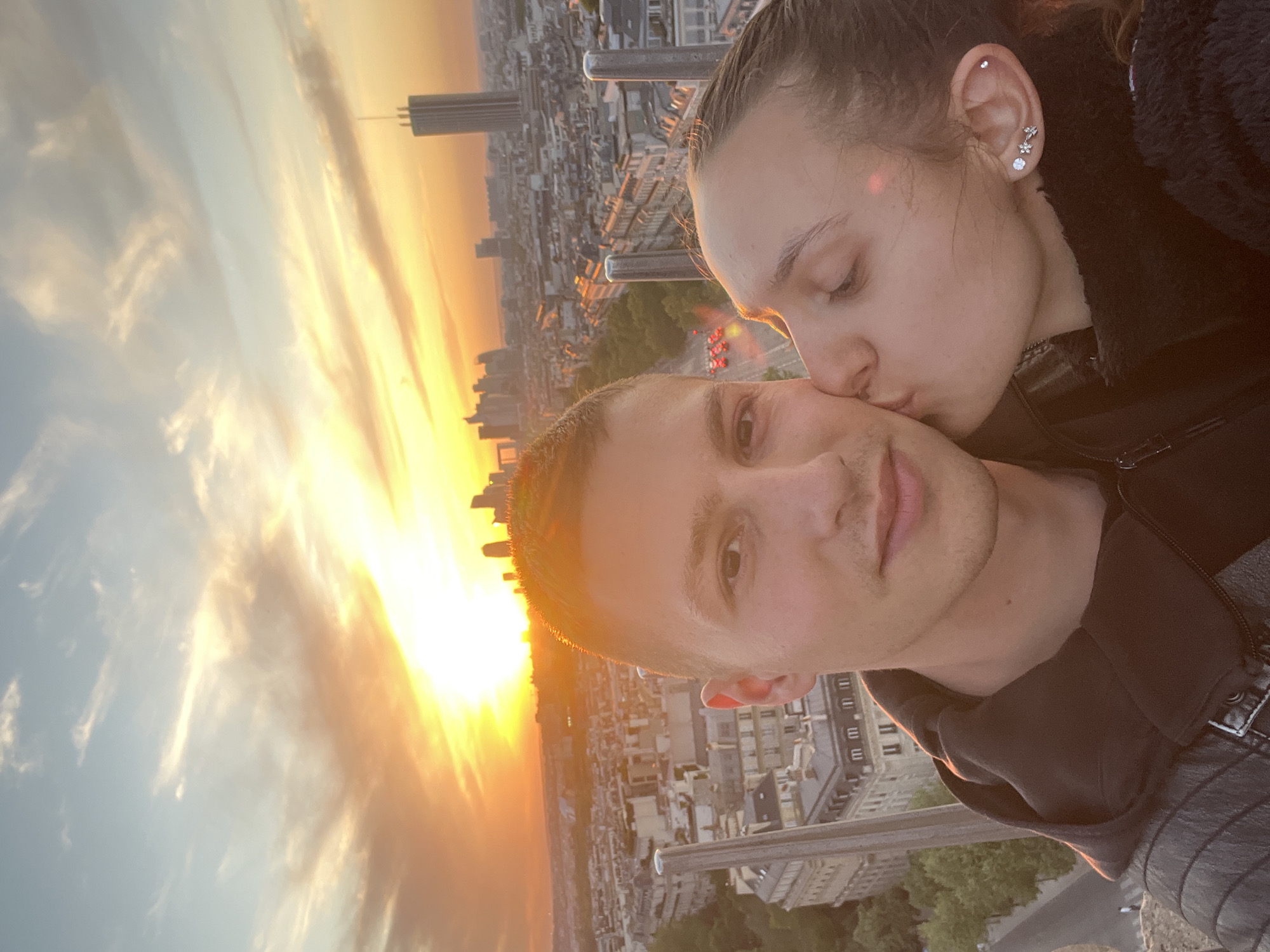An intimate portrait features a couple at sunset, captured from a high vantage point overlooking a city. The man, positioned slightly to the left of the frame, looks directly at the camera, while the woman, to his right, gently kisses his cheek with her eyes closed. She wears several distinctive earrings, including a stud at the top of her ear and a cluster of floral and larger stud designs lower down.
Behind them, a dramatic sunset dominates the sky, with the low sun casting a brilliant golden-orange glow across the horizon and illuminating scattered clouds. Below the sunset, a vast urban landscape stretches into the distance, filled with numerous buildings. A tall, dark, rectangular skyscraper, identifiable as the Montparnasse Tower, stands prominently in the background, confirming the location as Paris, France. The warm, golden hour light bathes the couple and the cityscape, highlighting their affectionate moment against the romantic backdrop of the setting sun over the French capital.