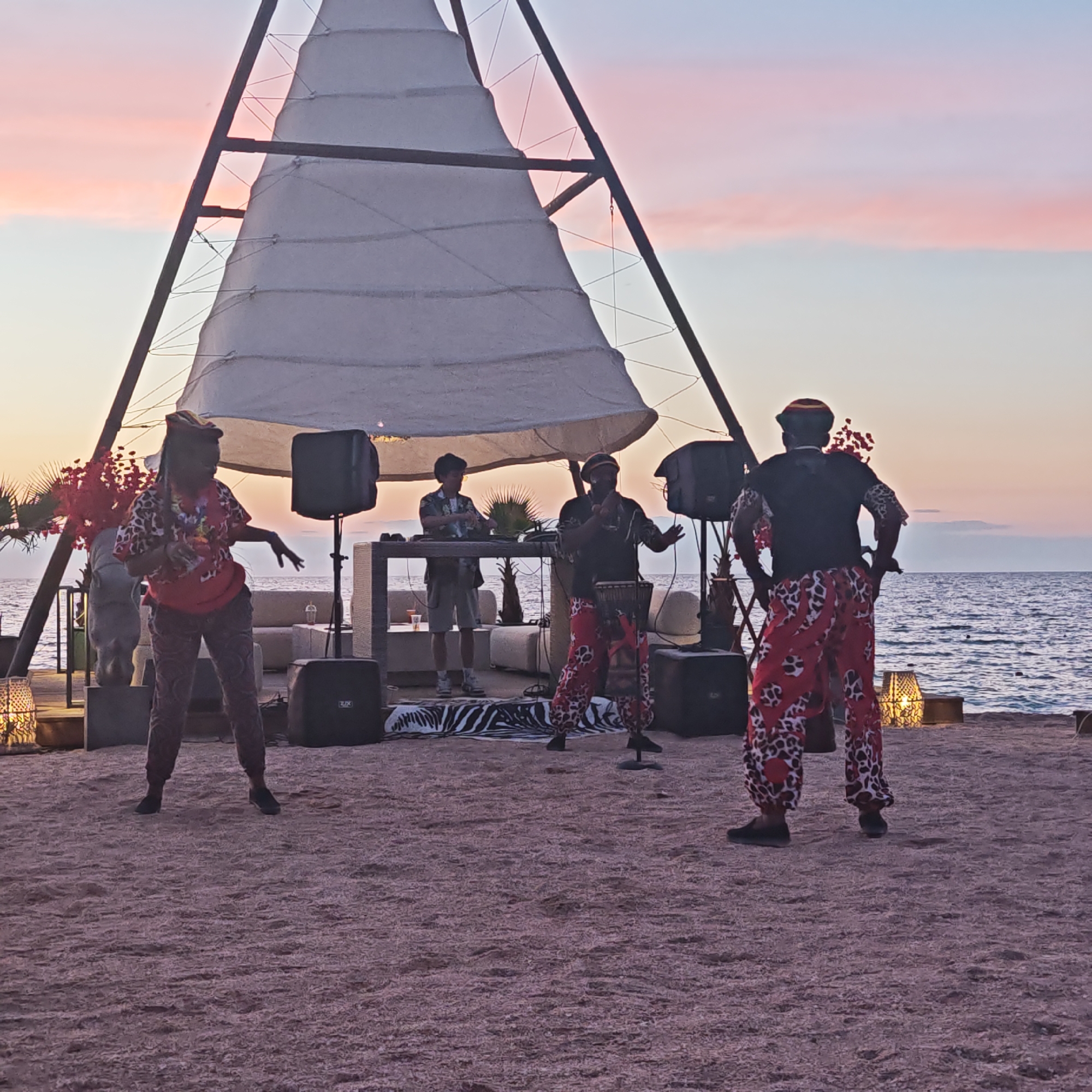 A group of four people are performing on a sandy beach at sunset. Two individuals in the foreground are dressed in vibrant, patterned clothing. One, on the left, wears a leopard-print shirt, red sash, and patterned pants, and appears to be dancing with hands outstretched. The other, on the right, faces away from the camera, wearing a black shirt with leopard-print accents and red patterned pants, with hands on hips. In the mid-ground, two other individuals are present. One, wearing a colorful patterned shirt and a hat, is playing a drum. To his left, another person in a patterned shirt and shorts stands near a DJ setup. Behind the performers, a large, white sail-like structure is supported by a wooden frame, resembling a triangular sail. Several black speakers are positioned around the performance area. The background features the calm sea and a sky with pastel hues of pink and blue, indicating sunset. The location is described as the city of Мысовское сельское поселение, Ukraine.