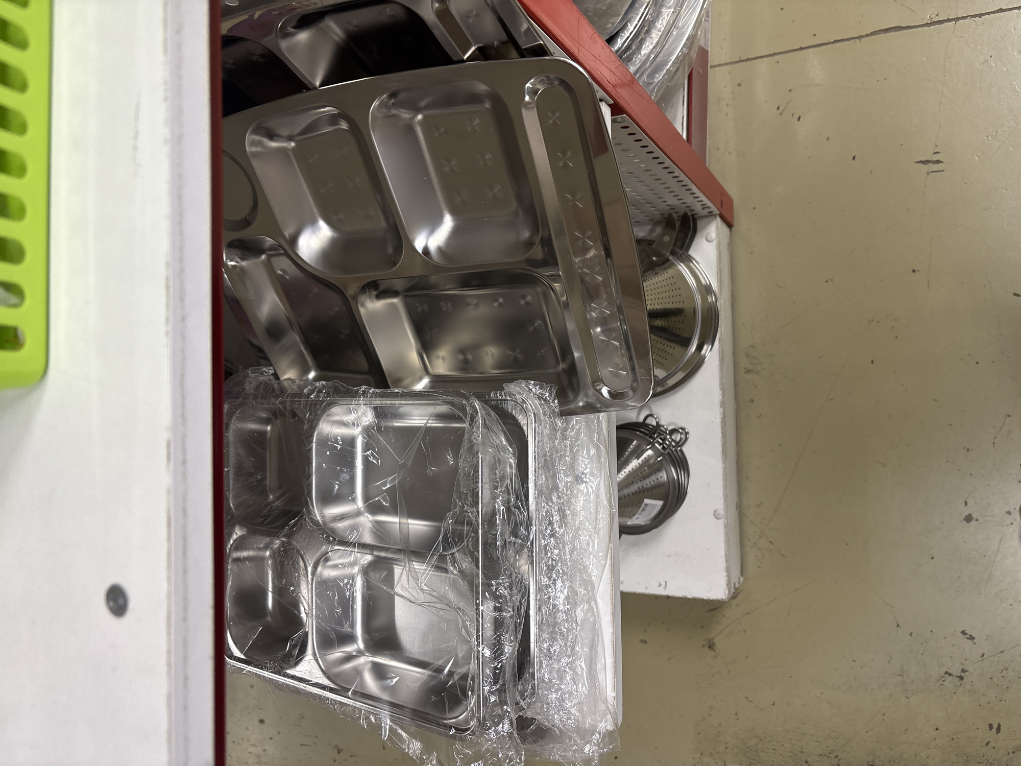 The image shows a shelf of stacked, stainless steel divided trays, likely for serving food in a cafeteria or institutional setting. Some of the trays are wrapped in plastic. Other kitchenware, possibly strainers, are also visible on the shelf. The image was taken indoors, likely in a storage area or warehouse. The floor appears to be concrete. No people are visible in the image. There is no indication that this image was taken in Krasnoyarsk, Russia.