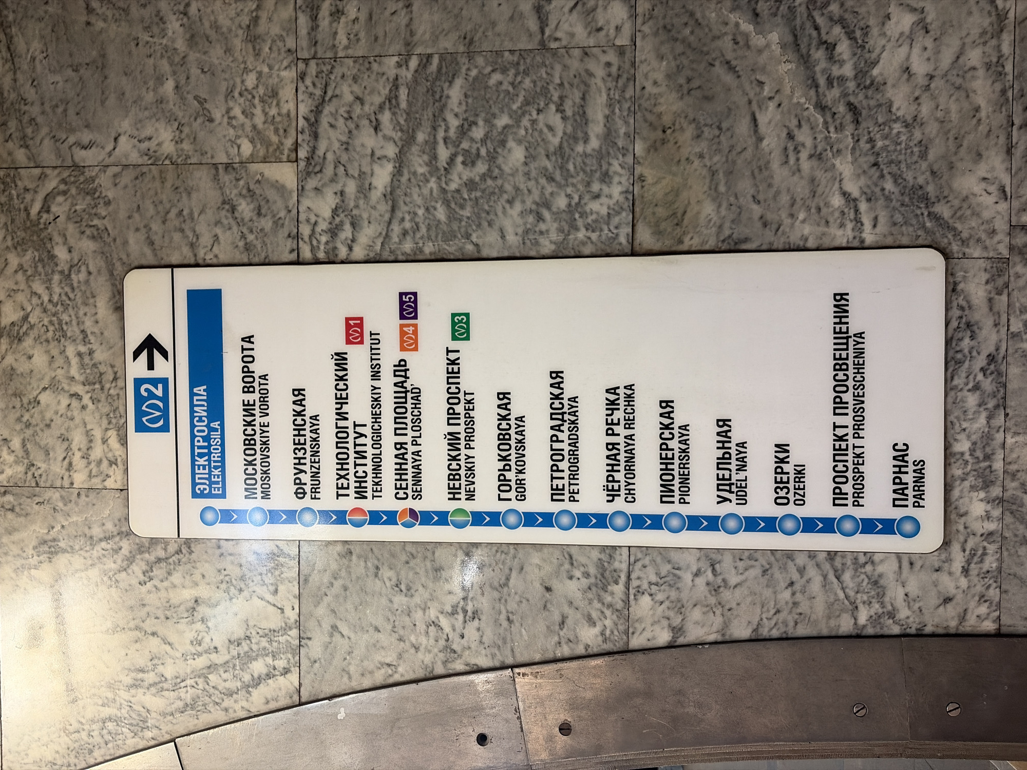 The image shows a sign from the Saint Petersburg Metro, Russia. The sign displays the stations on Line 2 (blue line) going towards Parnas. The stations listed include Elektrosila, Moskovskiye Vorota, Frunzenskaya, Tekhnologicheskiy Institut (with connection to Line 1), Sennaya Ploshchad' (with connections to Lines 4 and 5), Nevskiy Prospekt (with connection to Line 3), Gor'kovskaya, Petrogradskaya, Chyorna Recka, Pionerskaya, Udel'naya, Ozerki, Prospekt Prosvescheniya, and Parnas. The sign is mounted on a marble tiled wall or floor.