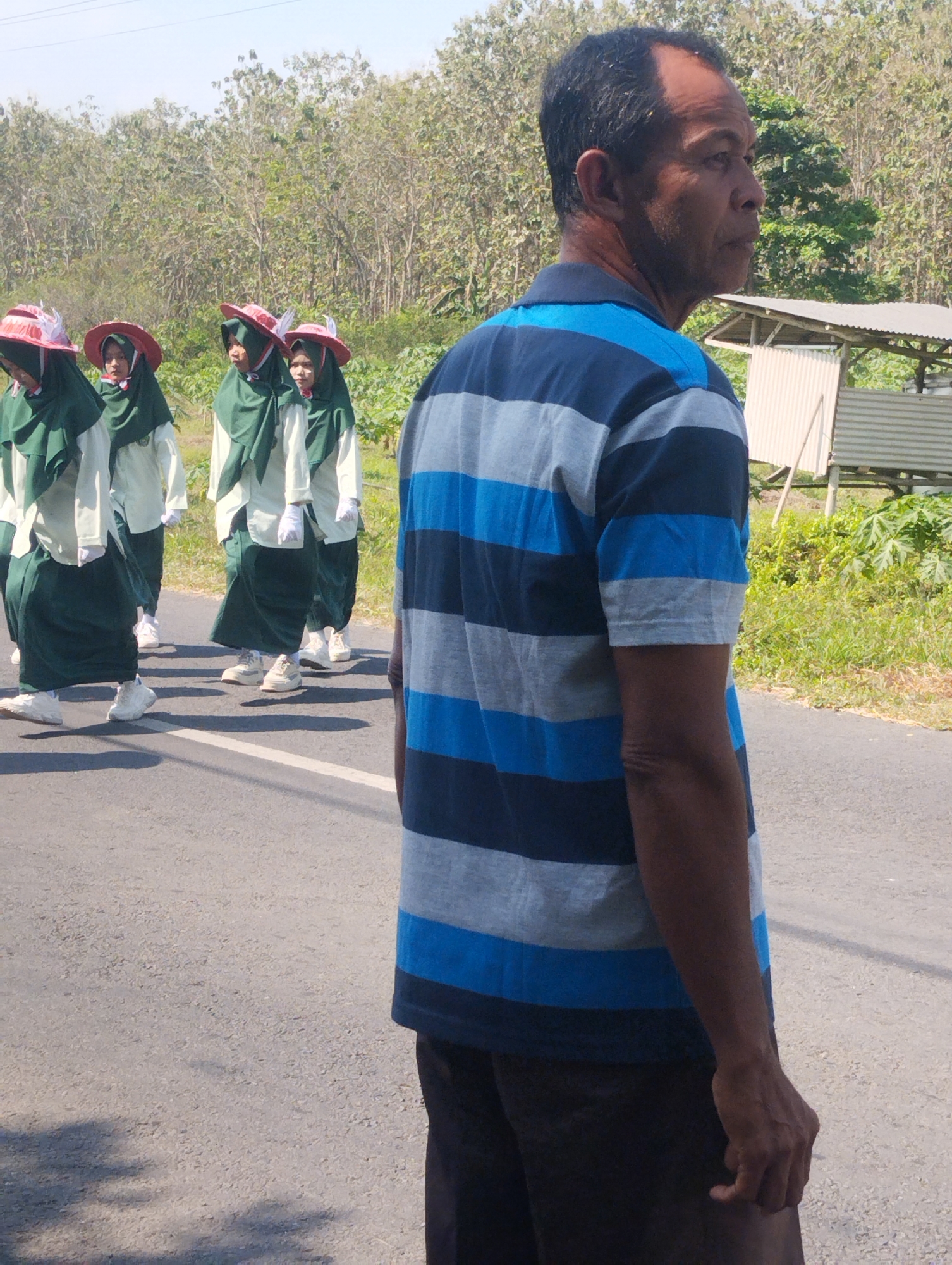 In Karangdoro, Indonesia, a man wearing a blue and gray striped polo shirt and dark pants stands on the right side of the frame, facing away from the camera and looking to the right. Behind him, a group of four young individuals wearing green hijabs and white shirts with dark green skirts are walking on a paved road. They are also wearing red hats with feather accents. The background consists of a line of trees and a simple, open-sided shelter with a corrugated metal roof. The sky is clear and blue, indicating a sunny day. Shadows on the road suggest the presence of trees or other objects casting them. The overall scene depicts a group participating in some form of procession or parade.