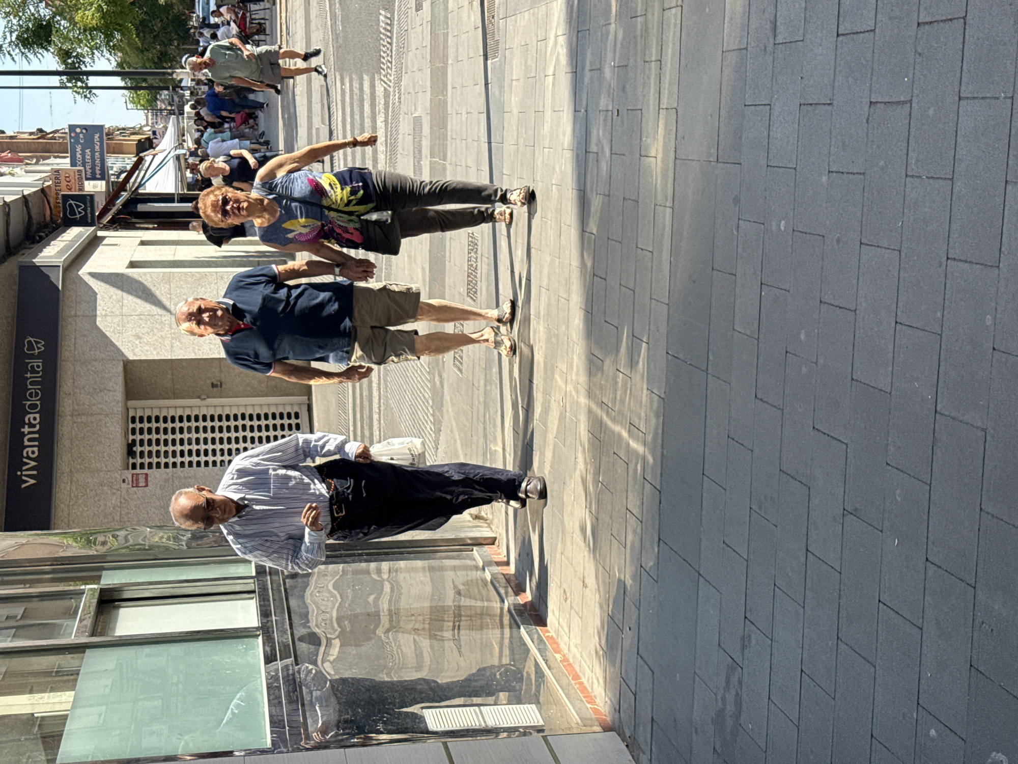 The image depicts a street scene. Three people are visible in the foreground: an older man in a light blue striped shirt and dark trousers, a middle-aged man in a dark blue polo shirt and khaki shorts, and an older woman in a multicolored top and dark trousers. The man in the striped shirt is walking in the opposite direction of the other two. There are more people further down the street. The street is paved with grey stones. A building is on the left side of the frame, with a sign reading "vivantadental." The location appears to be in the city of Unknown, Unknown.