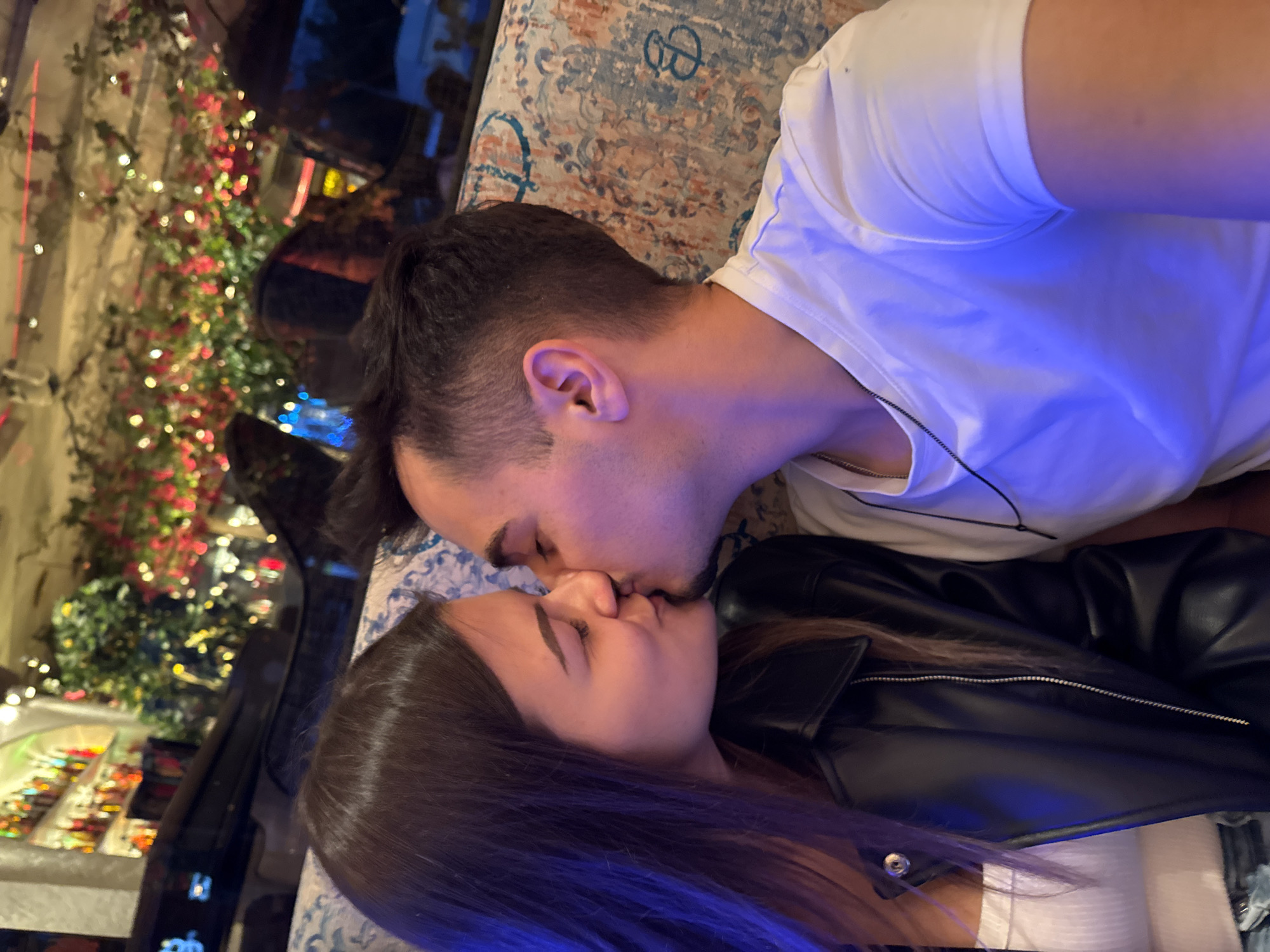 The image depicts a close-up profile view of two individuals, identified as a male and a female, kissing on the lips in what is identified as a restaurant or bar in the city of Sofia, Bulgaria. The female, positioned on the left, has long, straight dark brown hair and a fair complexion. She is wearing a white ribbed tank top underneath a black faux leather jacket. Her eyes are closed. The male, positioned on the right, has short dark hair and a fair complexion. He is wearing a white short-sleeved t-shirt and a silver chain necklace. His eyes are also closed. Both individuals are illuminated by a distinct blue light source originating from the left side of the frame, casting a blue hue on their faces, hair, and the male's shirt. They are seated on a patterned upholstered bench or sofa, which features an intricate design in blue, beige, and subtle orange tones, including repeated circular motifs resembling a stylized "B" or "D". The background is out of focus, revealing a dimly lit interior. Visible elements in the background include shelves stocked with various colored bottles, suggesting a bar area, and an overhead decoration of lush artificial foliage intertwined with numerous small, warm-toned fairy lights (red, yellow, and white), creating a festive ambiance. A large, dark, reflective surface, possibly a window or mirror, is also visible, reflecting some of the background lights.