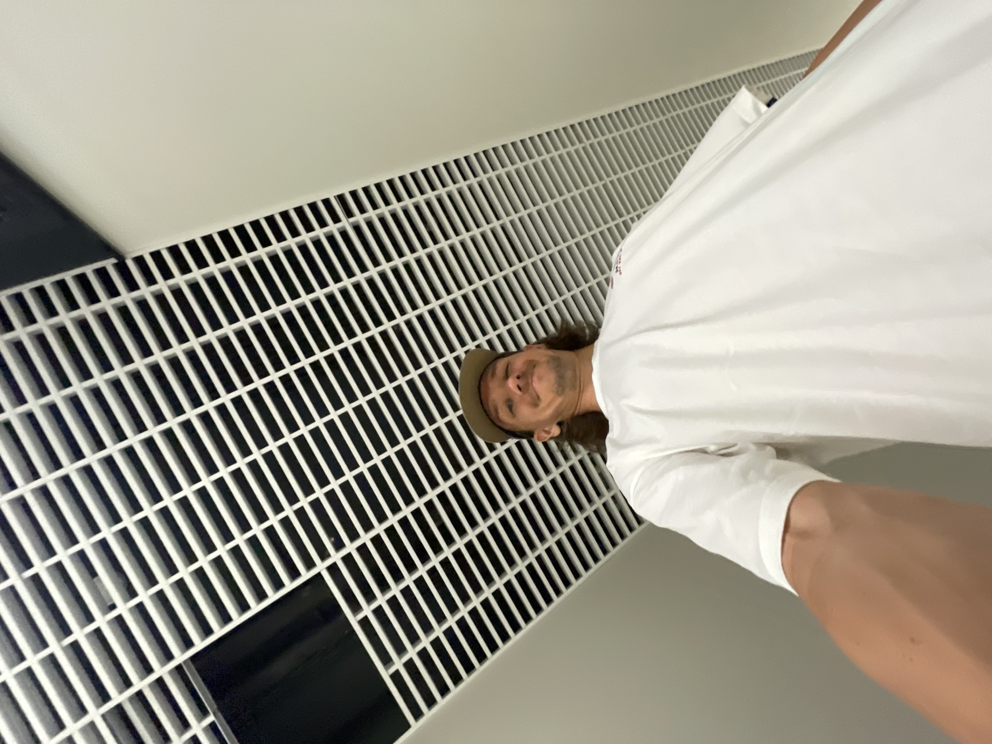 A man in a white shirt and tan hat is taking a selfie in front of a white, rectangular-gridded wall, possibly a vent. The wall is located near a black surface. The image is taken from a low angle looking up. No information indicates the location is in Izhevsk, Russia.