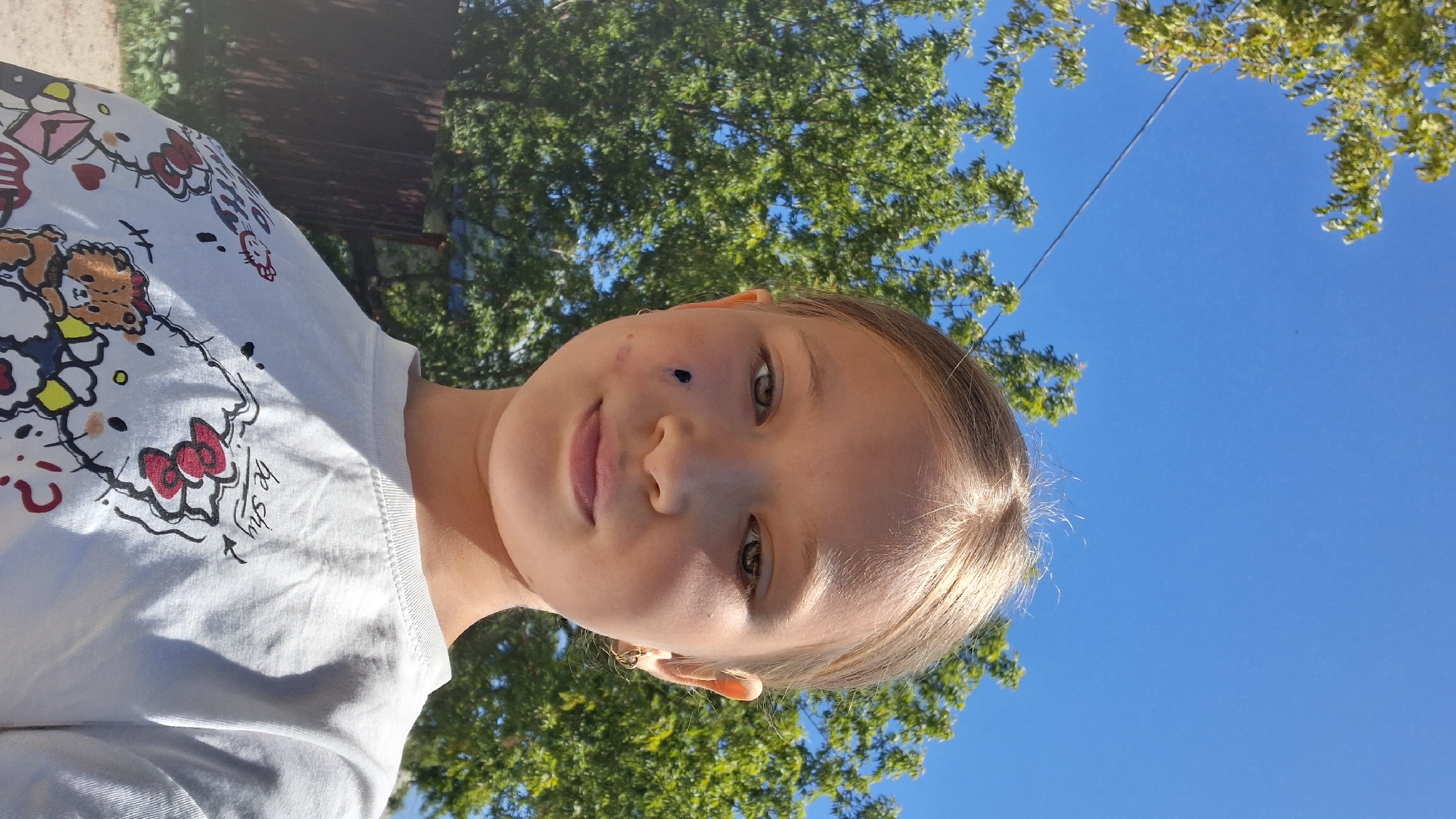 The media file is a portrait of a young girl taken outdoors. The girl is positioned in the center of the frame, looking directly at the camera with a slight smile. She has light brown hair and green eyes. There is a dark mark on her left cheek, close to her nose. She is wearing a white t-shirt with a cartoon print featuring Hello Kitty characters and a teddy bear. The print includes the text "be shy" in a handwritten font.
The background is a bright blue sky with green foliage from trees on the left and right sides, suggesting a sunny day. The angle of the shot, looking upwards, combined with the clear blue sky and sunlight, indicates it is daytime, likely mid-morning or afternoon. The presence of trees and open sky suggests a park, backyard, or similar outdoor recreational area. There are no other people visible in the frame. The overall mood appears cheerful and relaxed, characteristic of a casual outdoor photo. The location context provided is Semey, Kazakhstan.