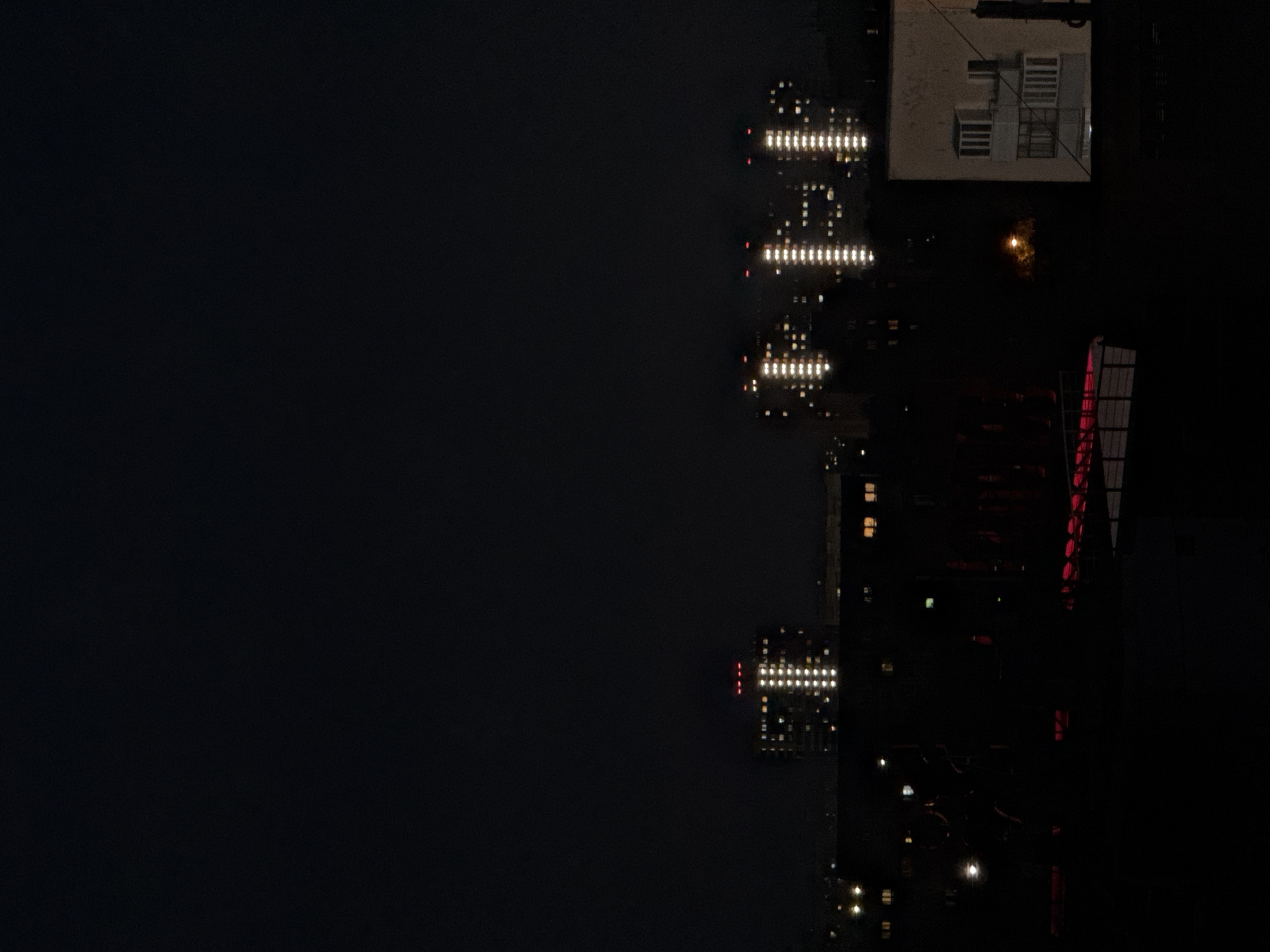 A nighttime cityscape in Nizhny Novgorod, Russia, is depicted from a high vantage point. The sky is completely dark, suggesting it is after sunset or before sunrise. In the background, three tall apartment buildings are illuminated by rows of bright white lights, indicating that many apartments are occupied. Each of these buildings also features red lights on their uppermost sections. In the mid-ground and foreground, the scene is largely obscured by darkness, but faint outlines of other buildings and structures are visible. One building in the lower right corner of the frame is partially illuminated, revealing multiple windows and a balcony structure. A bright yellow light, possibly from a streetlamp or a distant illuminated area, is visible in the middle distance amidst darker foliage. Horizontal red lighting is present in the lower middle section of the image, possibly from decorative lights or a sports facility. The overall impression is of a sprawling urban environment at night, with residential buildings standing out against the darkness.
