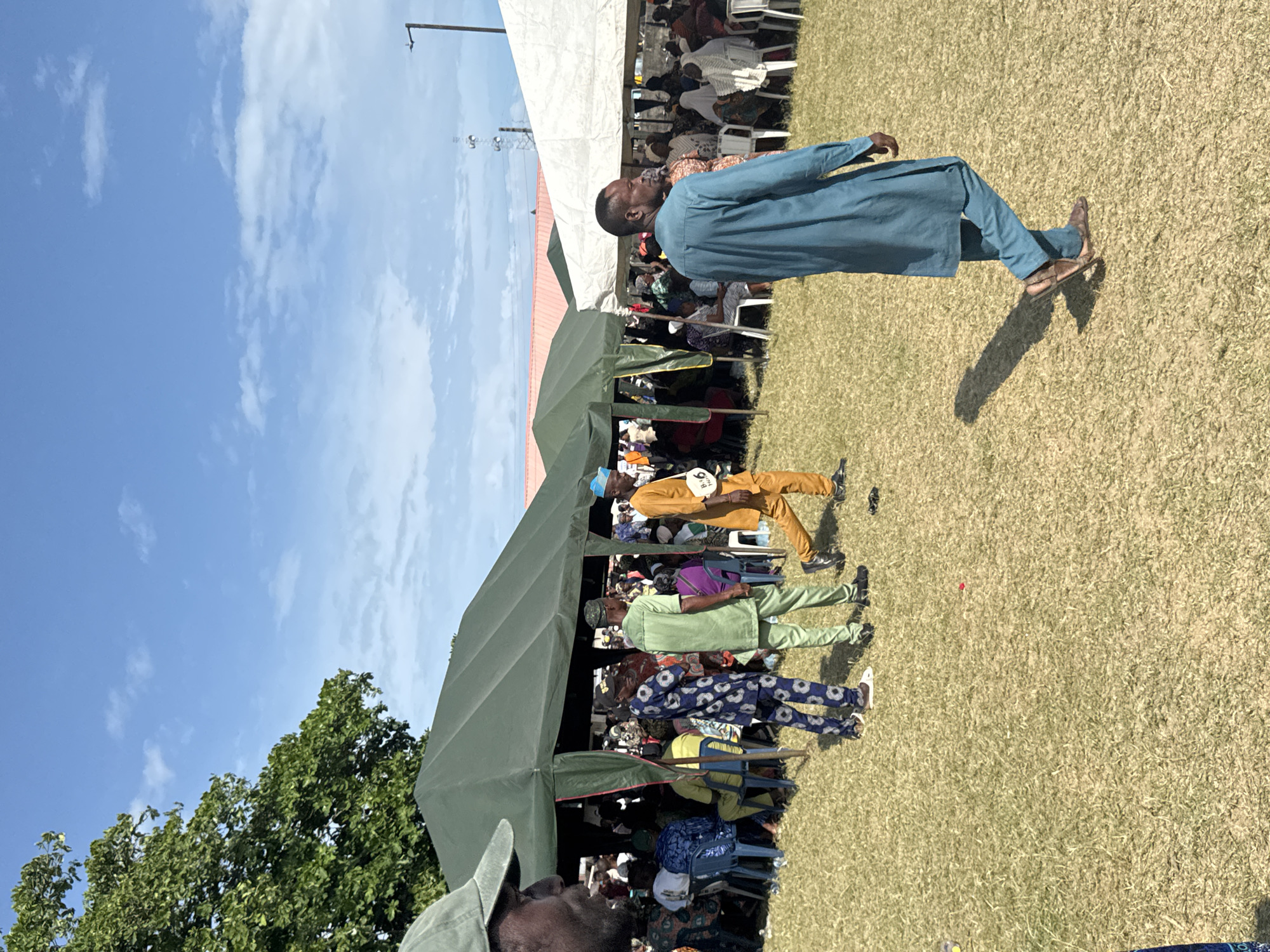 The image shows an outdoor gathering in Badagry, Nigeria. The scene is populated by a large crowd, some seated in rows of white plastic chairs under a white tent and a green tent. Several people, dressed in traditional African attire, are standing and walking near the tents. One man, wearing a blue outfit, stands apart, gazing towards the crowd. The backdrop features a clear blue sky with scattered clouds and some green trees. The ground is covered in short-cut grass.