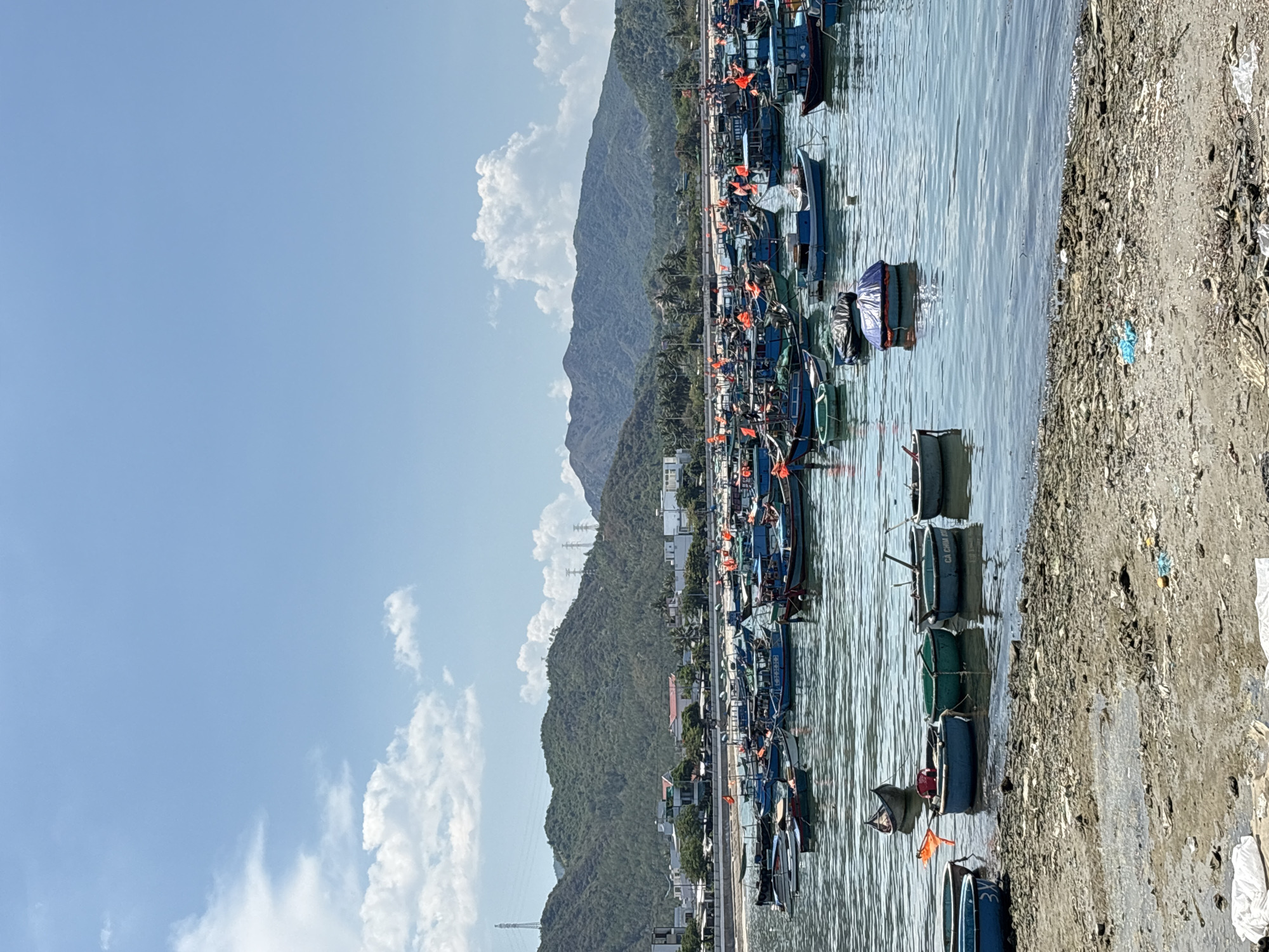The image depicts a coastal scene in Vĩnh Lương, Vietnam. A multitude of small boats are moored in the calm water along the shoreline. The boats vary in color and design, with several displaying orange flags. A mountain range is visible in the background, adjacent to a scattering of buildings and houses. The sky is pale blue with scattered white clouds. A concrete shore with litter and debris runs along the water’s edge. No people are clearly visible.