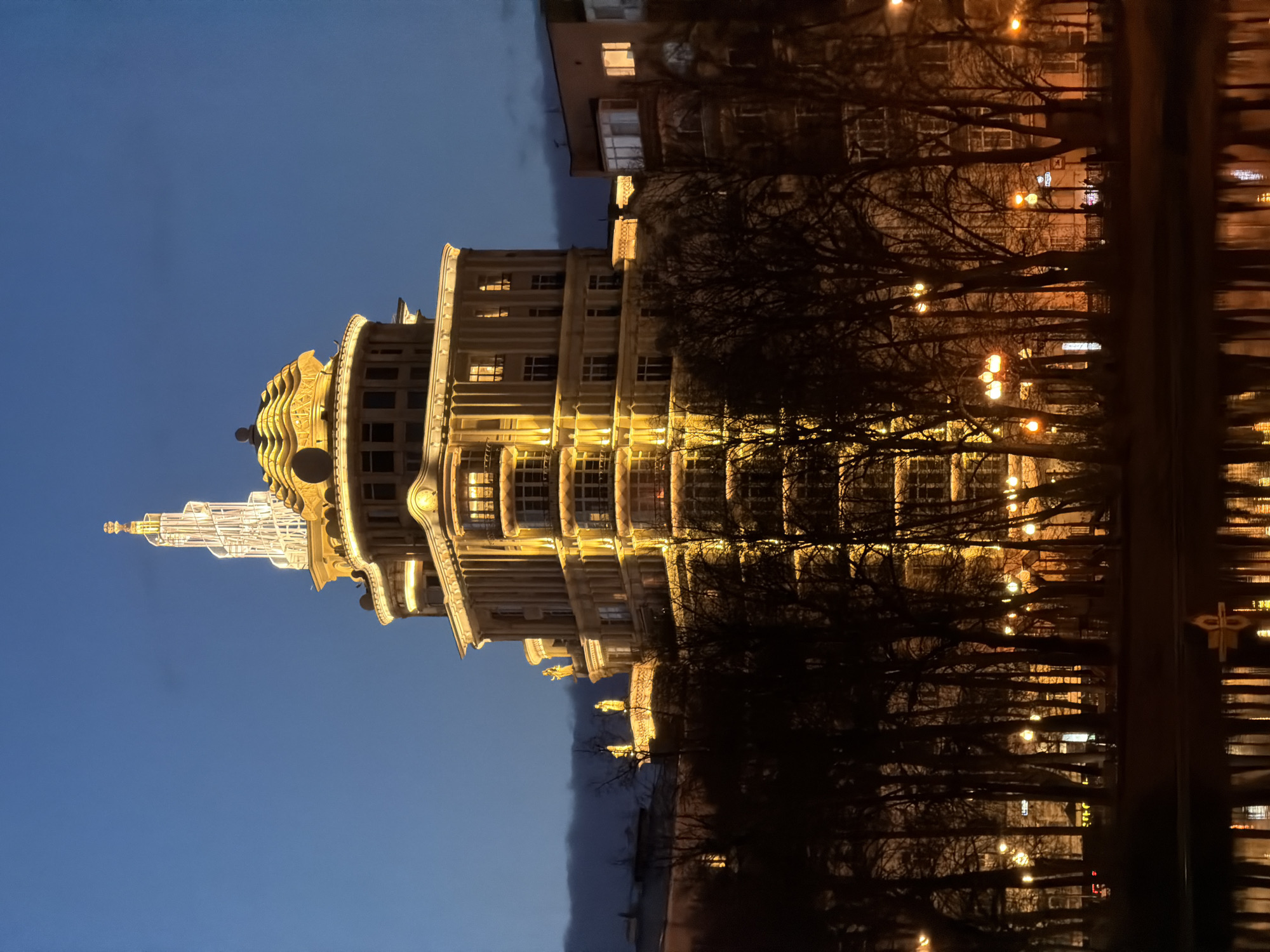 The image depicts a nighttime view of the Kudrinskaya Square Building, one of the "Seven Sisters" skyscrapers in Moscow, Russia. The building is illuminated with warm lighting, highlighting its architectural details. To the right of the building is a park or street lined with trees that are mostly bare, suggesting it is late autumn or winter. Streetlights are visible through the trees. The sky is a gradient from dark blue to black. Reflections of the building and lights are seen in a dark, possibly wet surface, adding to the nighttime atmosphere. Another building is seen directly next to the Kudrinskaya Square Building on the right. There are no visible people in the image.