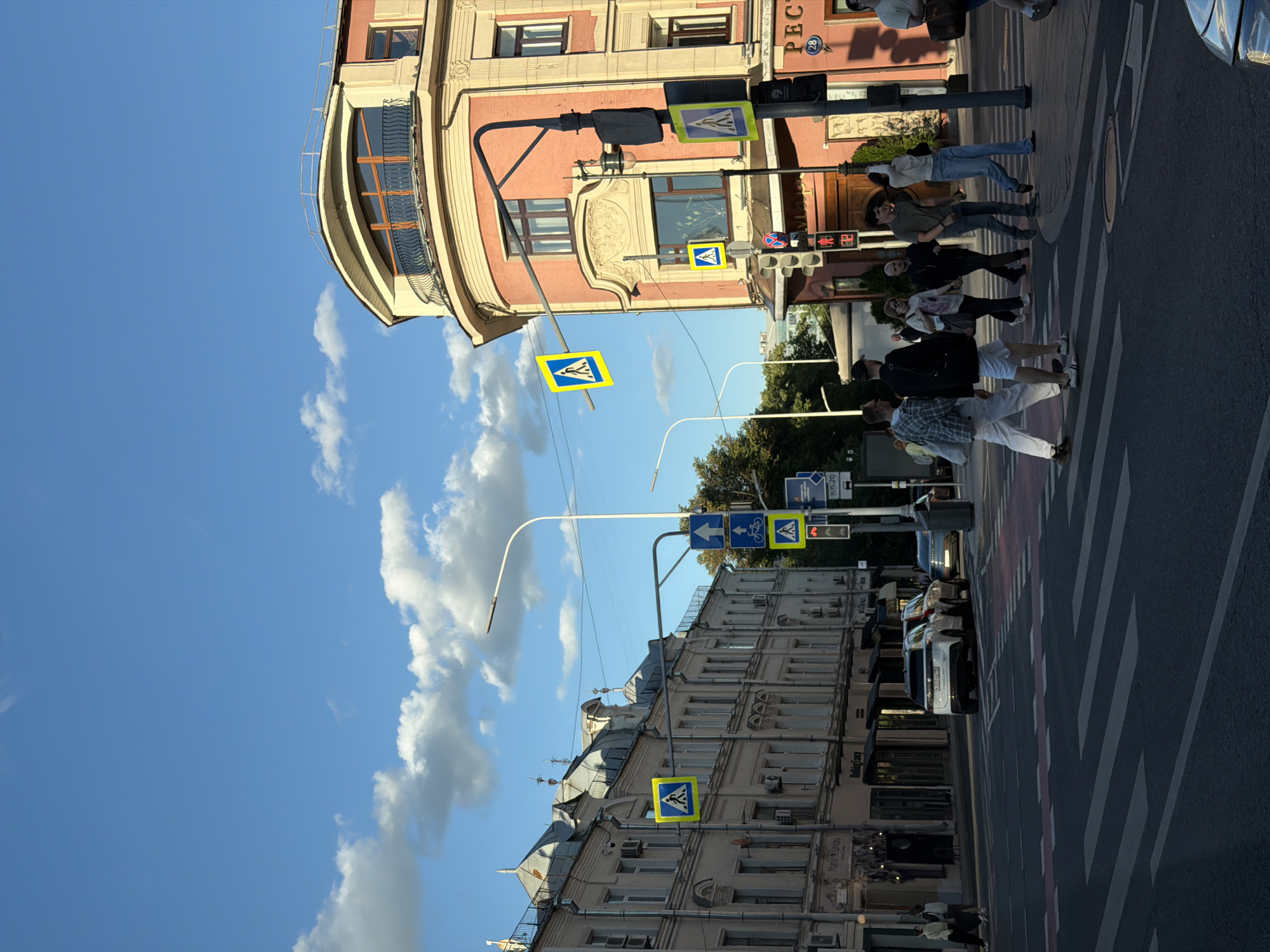 This is a vertical, eye-level shot of a street scene in Moscow, Russia, under a bright blue sky with scattered white clouds. The image is oriented with the top of the frame to the left.
The foreground shows a crosswalk with several people walking across it. The people are diverse in age and attire, some appearing to be tourists, others locals. They are crossing from right to left in the image's orientation.
The middle ground features buildings on both sides of the street. The building on the left is a prominent, older structure with a curved, decorative facade and several windows. The building on the right is also an older building with a more ornate facade, featuring detailed stonework and multiple windows. Traffic lights and pedestrian crossing signs are visible on poles. The pedestrian signs are yellow squares with a blue symbol of people walking.
The background includes more buildings and trees, suggesting an urban environment. Several cars are parked or driving on the street, including a white SUV visible on the right side of the crosswalk.
The lighting indicates it is daytime, likely late afternoon, given the warm light and shadows. The weather appears clear and pleasant.
Visible text includes a sign with "РЕСТ" written in Cyrillic on the pink building. There are also numbers visible on some traffic signs, such as "28" and what appear to be traffic signal timings.