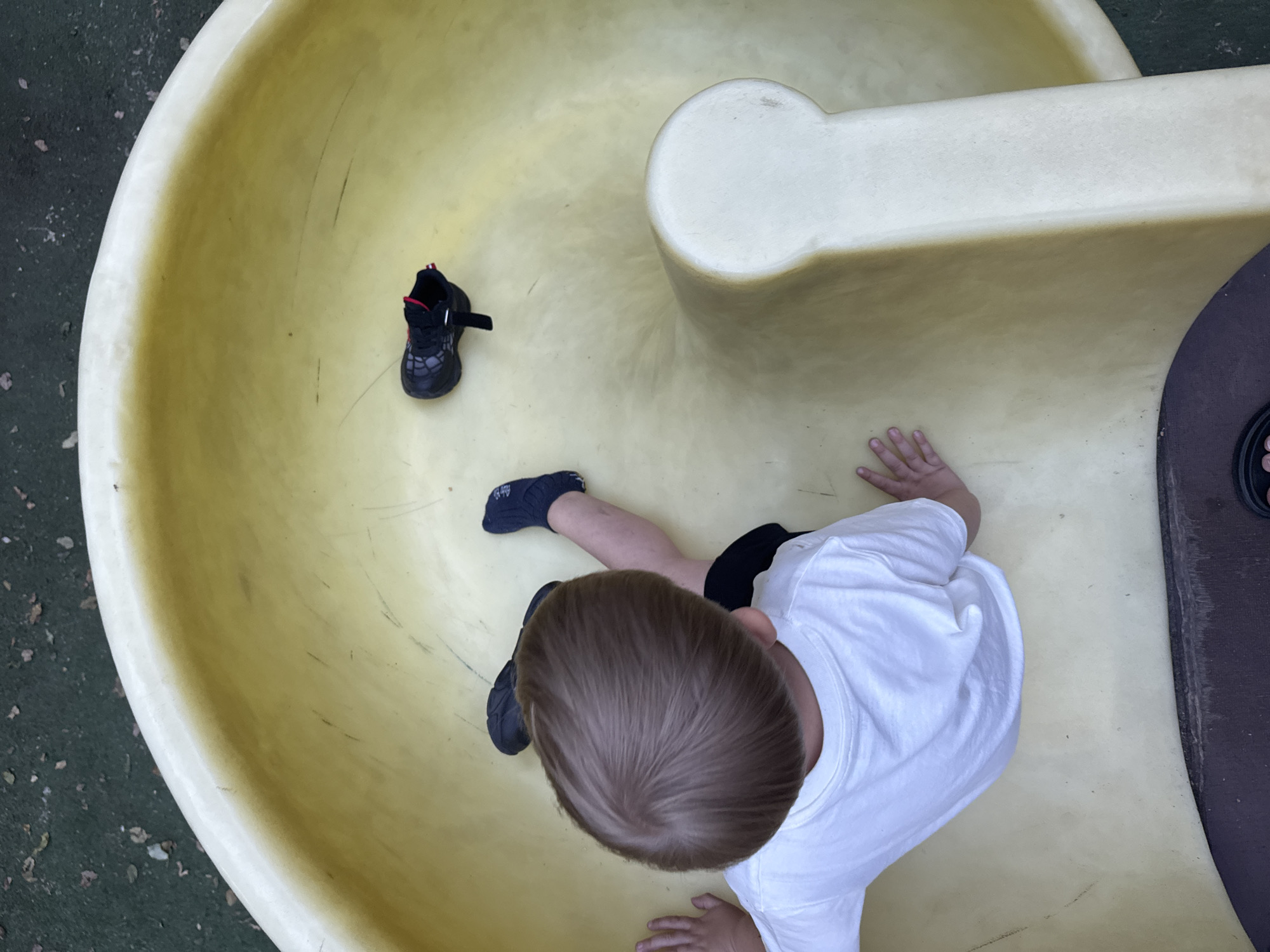 This is a top-down, close-up, medium shot of a young child sitting on a yellow plastic slide. The child is wearing a white t-shirt and black shorts, with one black shoe on their left foot and the right shoe is off. The slide is curved, with a white handrail at the top. The ground around the slide appears to be dark green mulch with some scattered leaves.
The child's hair is light brown and short. Their left hand is resting on the slide near their leg, and their right hand is visible near the edge of the slide, suggesting they are preparing to slide or have just finished. The child's expression is not visible from this angle. The lighting suggests it is daytime, possibly a sunny day, given the bright colors and clear visibility. The overall setting appears to be an outdoor playground. There is no visible text in the image.