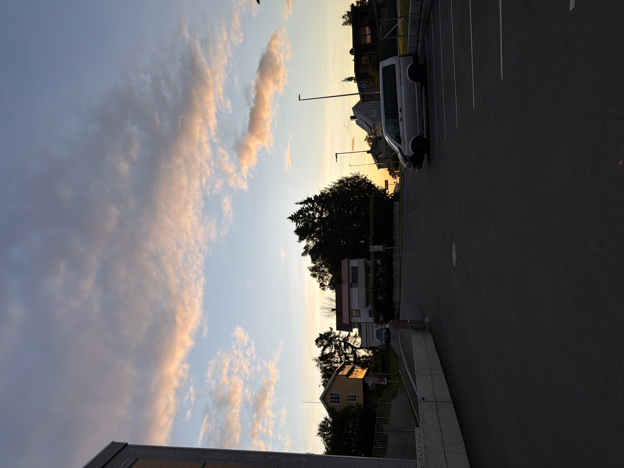 An exterior, medium-shot photograph captures a parking lot with a silver van in the foreground. The van is parked in a marked parking space. In the background, several houses and trees are visible against a sky with clouds tinged with orange and pink hues, suggesting either sunrise or sunset. A building with a corrugated facade is on the left edge of the frame, and a concrete barrier runs along the left side of the parking lot. Streetlights are visible in the middle ground, and a road slopes upwards towards the houses. The scene is located in the city of Harstad, Norway.