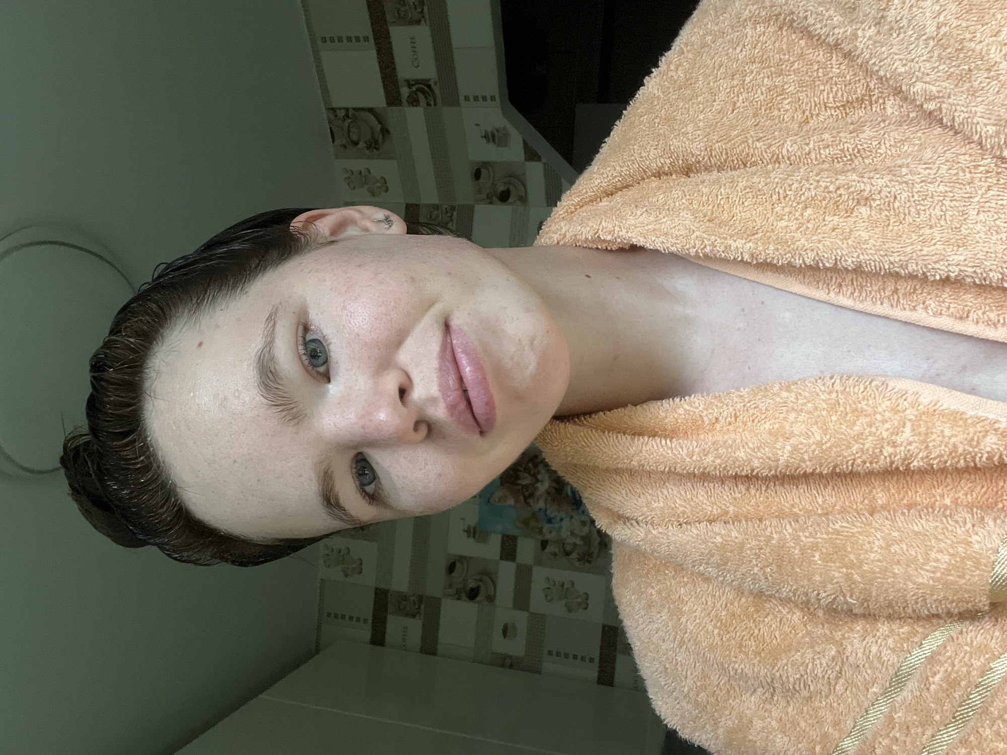 A young woman with dark hair pulled into a bun and blue eyes is wearing a peach-colored bathrobe. She is looking directly at the camera with a slight smile. Her skin is fair with some freckles. The background appears to be a tiled kitchen or bathroom with a coffee cup motif. A light fixture is visible on the ceiling. The overall lighting is soft and natural. The image was taken in the city of Chaikovsky, Russia.