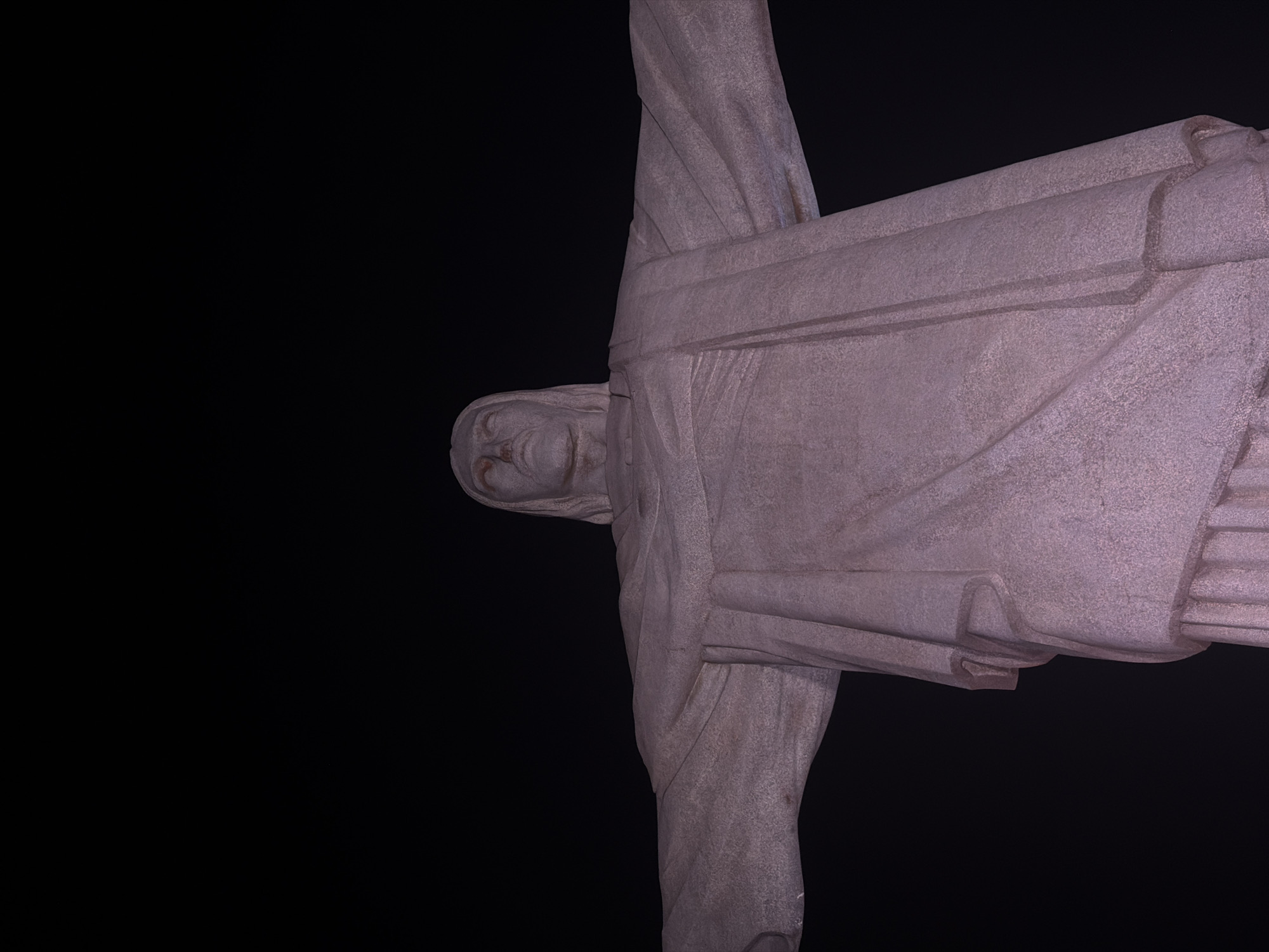 A low-angle, full shot of the statue of Christ the Redeemer is captured against a black background. The statue is made of stone and appears to be lit from below, casting a soft pinkish-purple hue. The statue's arms are outstretched in a gesture of welcome. The face of Christ is visible, with eyes looking downwards. The fabric of the robe is detailed with sculpted folds and lines. The statue stands on Corcovado Mountain in Rio de Janeiro, Brazil.