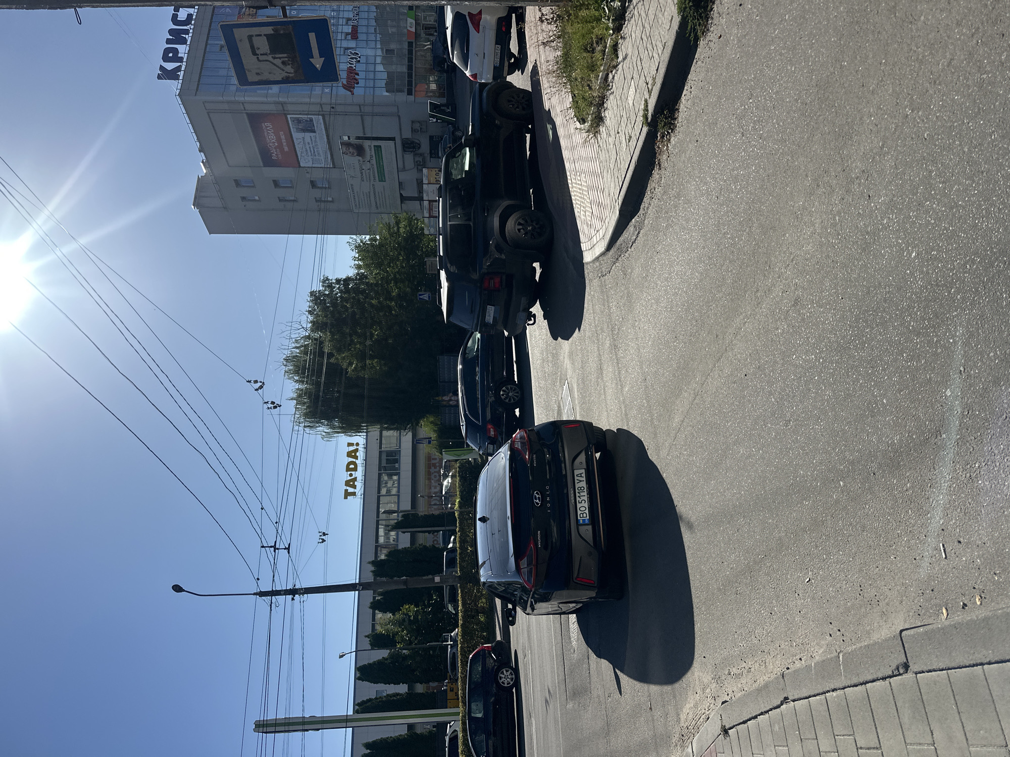The image depicts a street scene in Ternopil, Ukraine, on a sunny day. In the foreground, a black Hyundai Ioniq electric car with license plate BO 5118 YA is parked on the right side of the road, casting a shadow. A dark-colored SUV is also visible in the middle ground, positioned to the right of the Ioniq. Several other cars are parked or driving in the background.
To the right of the road, a building with "КРИСТ" (Krist) in large letters on top is prominent. This building appears to be a commercial establishment, possibly a shopping center or office building, as evidenced by signage such as "TA-DA!" and other advertisements. A blue directional sign with a gas pump icon and an arrow pointing left is mounted on a pole near the building.
The environment features a clear blue sky, power lines crisscrossing overhead, and trees lining the sides of the street and behind the buildings. The street is paved with asphalt, and a sidewalk made of grey rectangular bricks is visible on the left side of the image. The sun is shining brightly, creating lens flare in the upper right portion of the image. The scene appears to be an intersection or a street with moderate traffic.