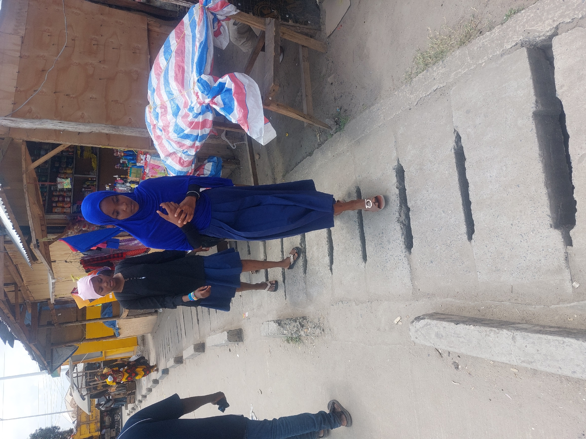 Two young women stand on a concrete sidewalk alongside an open market. The first woman wears a bright blue hijab, a blue top, and a long blue skirt. The second wears a light pink head covering, a black long-sleeved shirt, and a short blue skirt. Both wear sandals. The market stalls behind them display various wares, including canned goods, clothing, and household items. A red, white, and blue striped tarpaulin is draped over one of the stalls. Other people are visible in the distance near the market stalls. A portion of a person's legs and feet, wearing jeans and brown shoes, are visible at the bottom of the frame. The location is Dar es-Salaam, Tanzania.