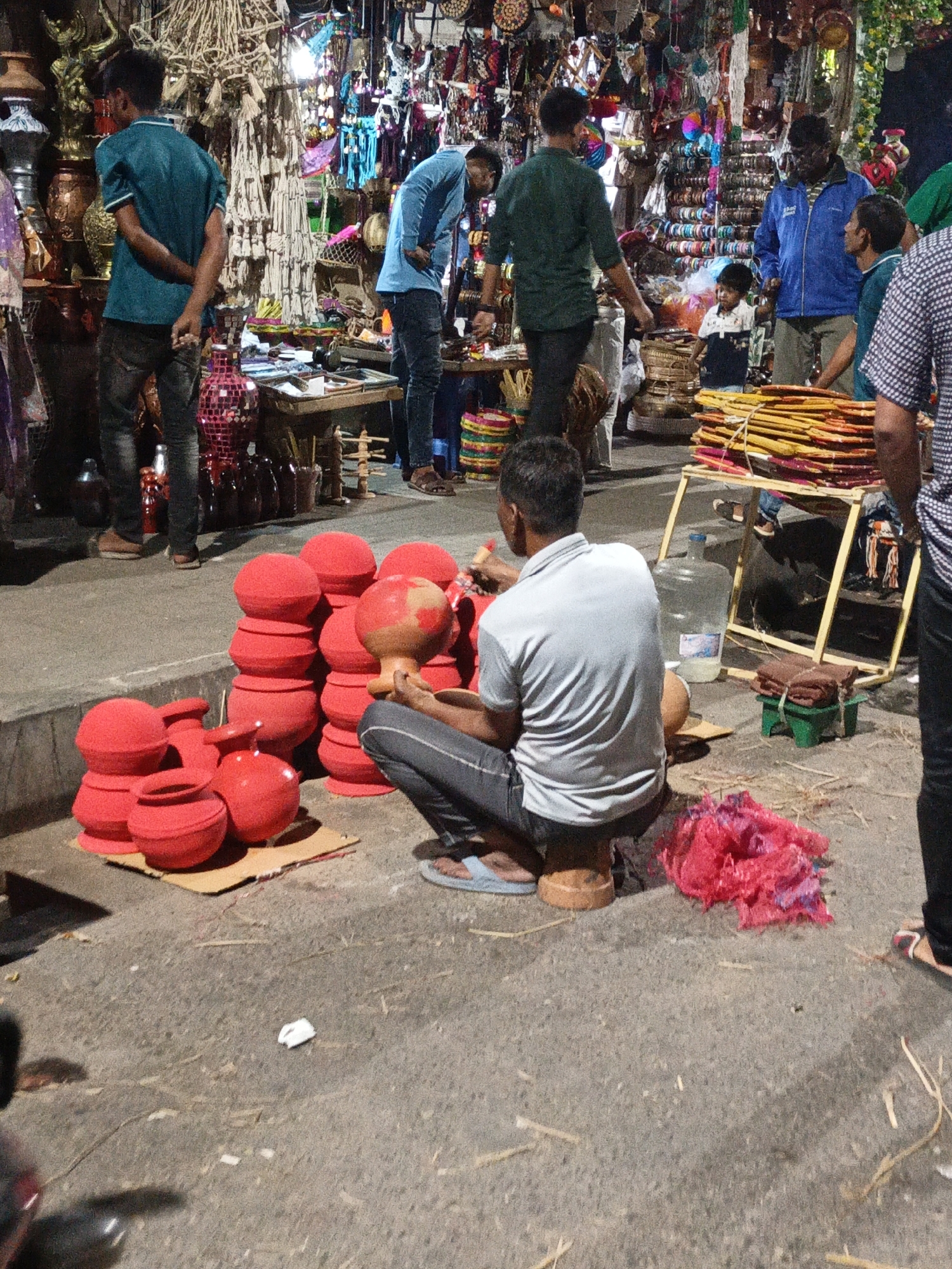 The image shows a street scene, likely in Dhaka, Bangladesh, based on the dense market environment and the products sold. A man sits on a small wooden stool, painting a clay pot with red paint, surrounded by stacks of already painted pots. He wears a grey shirt and dark pants, with blue sandals. A plastic water jug and a small green stool are nearby. A discarded piece of red fabric lies on the ground next to him. In the background, a bustling market scene unfolds with people browsing stalls filled with various handicrafts, decorations, and other items. Several men are seen examining the merchandise. The market is crowded, with a variety of colors and textures visible in the background. The location appears to be outdoors, on a paved surface.