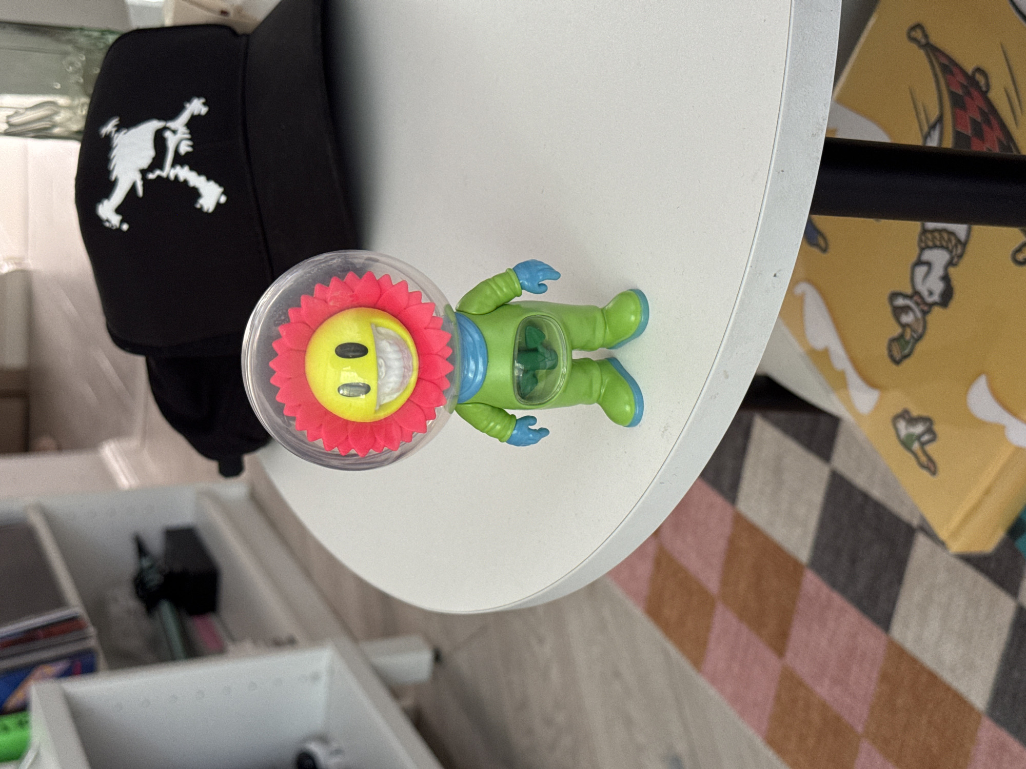 A close-up, slightly rotated view shows a white, round table with a black baseball cap, a toy astronaut with a flower head, a yellow and black checkered pillow with Snoopy and Woodstock cartoon characters, and a multi-colored checkered rug. The baseball cap is black with a white logo. The astronaut toy has a clear helmet with a yellow smiley face surrounded by red petals, a green body, and blue gloves. The table surface is clean and reflects light. A white storage organizer with various items is visible in the background. The location is unspecified and not identifiable as Moscow, Russia from visual elements.