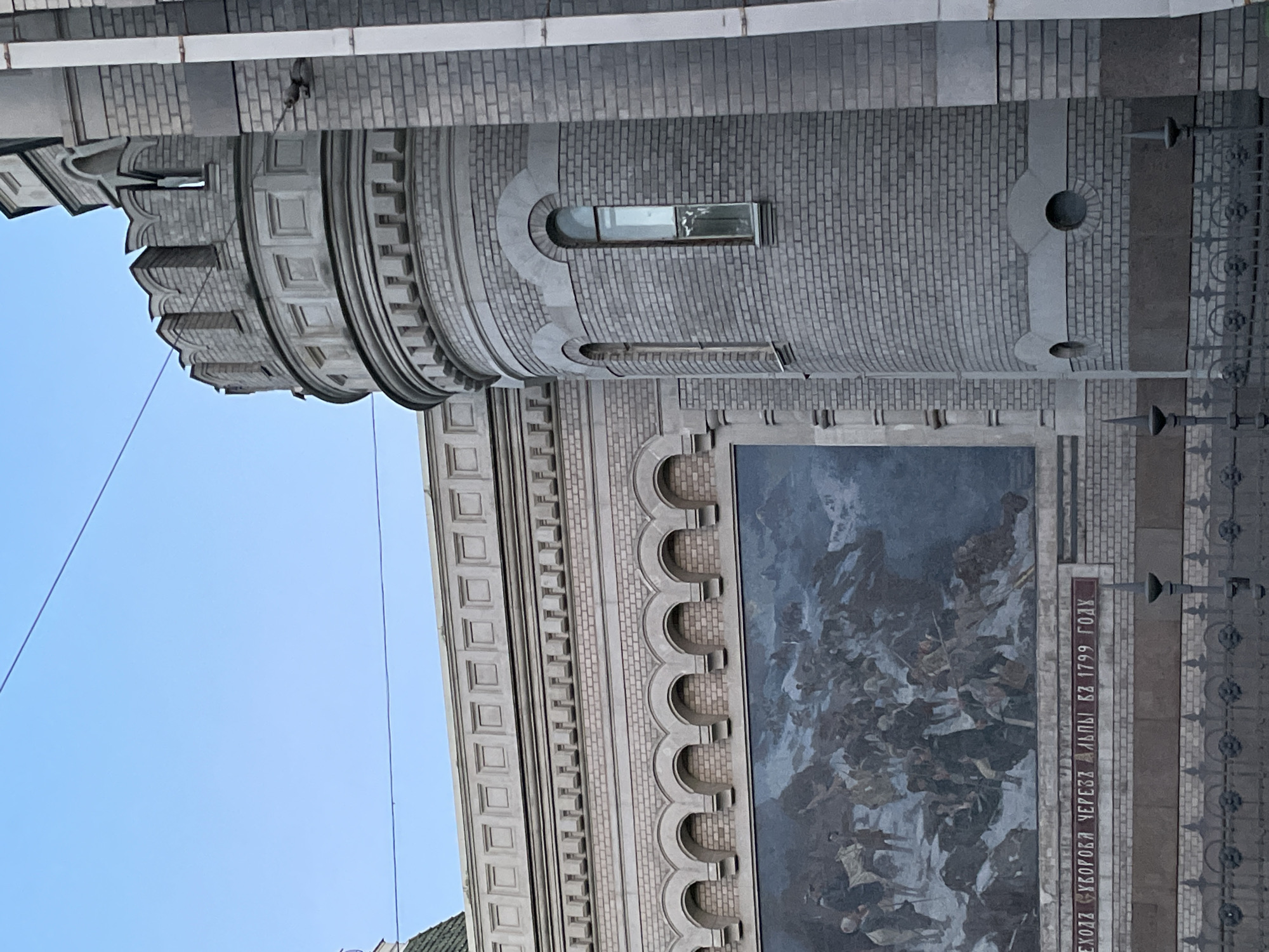 The image shows a building in Saint Petersburg, Russia, with a mural depicting a historical scene of a military crossing of the Alps. The building features a prominent round tower with battlements and arched windows, constructed of light grey brick. Adjacent to the tower is a long, flat section of the building with a decorative frieze and a series of arched alcoves. The mural, set within these alcoves, shows a large group of figures on horseback and on foot navigating a snowy, mountainous terrain. Below the mural, a red plaque displays text in Russian, which reads "ПЕРЕХОД СУВОРОВА ЧЕРЕЗ АЛЬПЫ В 1799 ГОДУ," translating to "Suvorov's Crossing of the Alps in 1799." The scene is framed by a dark metal fence in the foreground. The sky above is a clear, pale blue.
