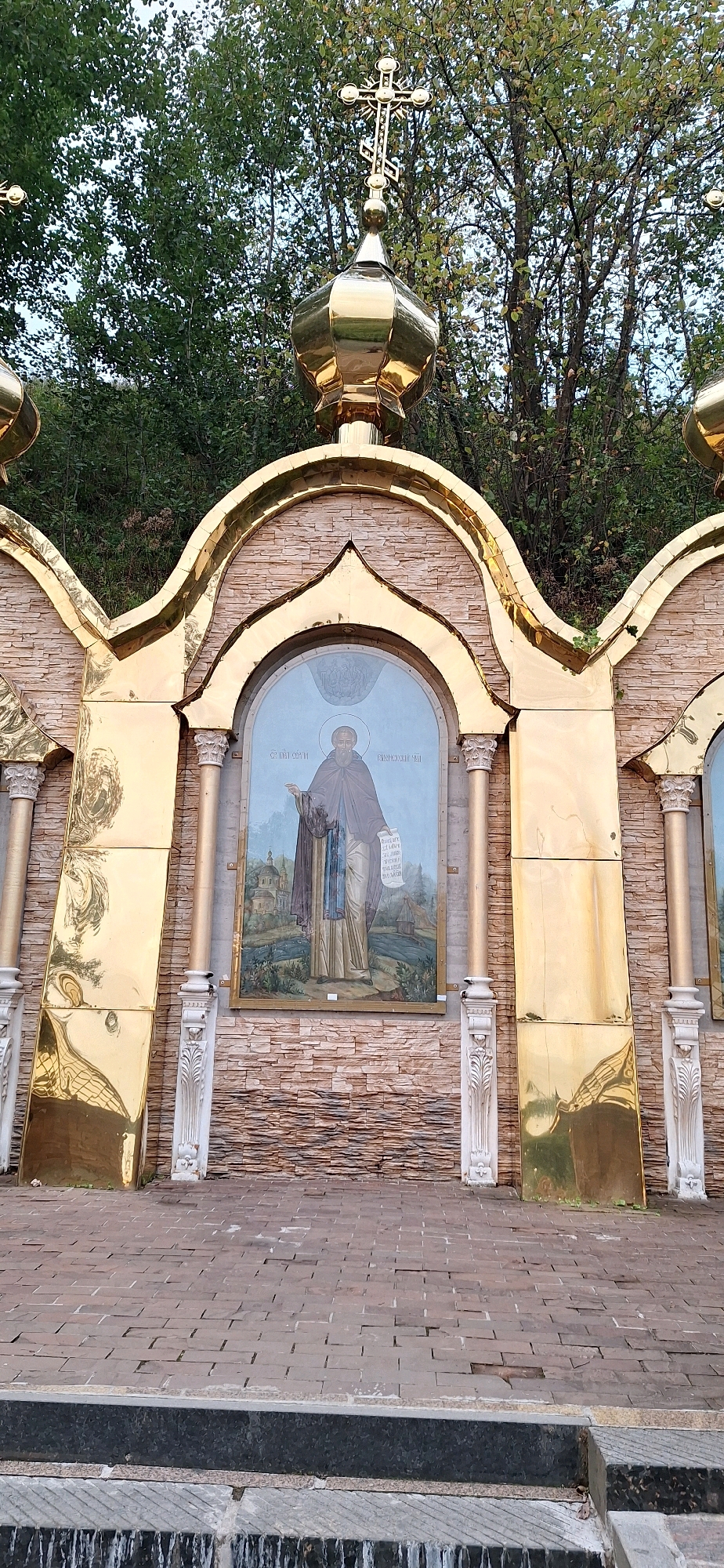 A frontal, eye-level shot shows a gold-colored dome with a cross topping an arched structure made of golden, reflective material and light brown stone tiles. Beneath the dome, within an arched alcove, is a framed religious painting depicting a man in religious attire holding a scroll. The painting is set against a backdrop of trees. The structure stands on a paved area of interlocking stones, with a dark gray stone step and a fountain in the foreground. This scene is located at the Holy Spring of St. Sergius of Radonezh in Radonezh, Russia.