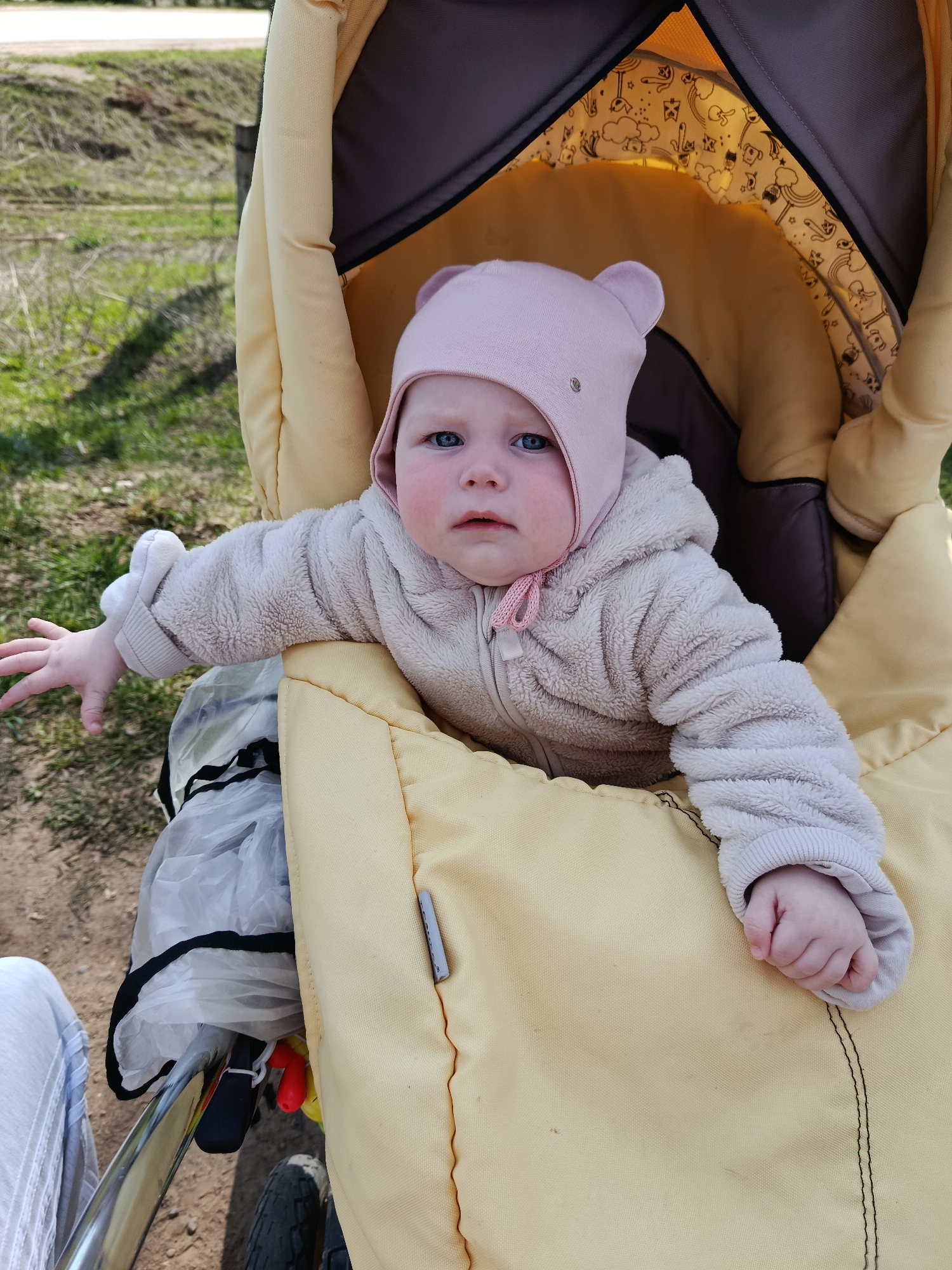 A baby sits in a yellow stroller, wearing a pink hat with bear ears and a beige fuzzy jacket. The stroller has a brown and patterned interior. The baby has blue eyes and is looking directly at the camera with a slightly concerned expression. The baby's left arm is outstretched. Outside the stroller, there is a glimpse of green grass and brown earth. A portion of a person's leg is visible on the left side of the image. The location is presumed to be in the city of Фоки, Russia.