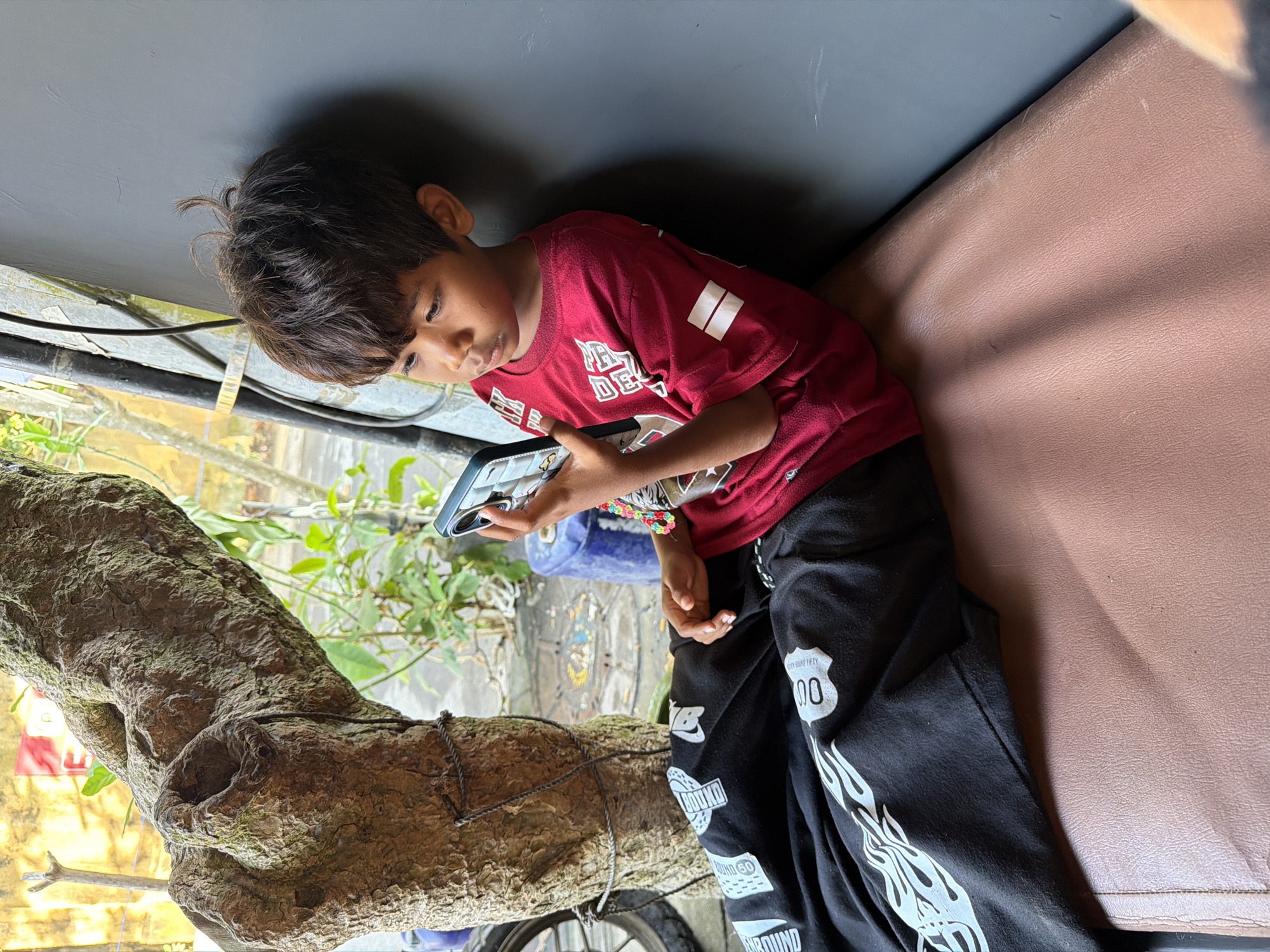 A young boy sits on a brown upholstered bench next to a gray wall, looking at his phone. He is wearing a red t-shirt with white graphics and black pants with white flame and "00" graphics. A thick tree trunk is to his left, and a window with greenery outside is visible. The scene appears to be outdoors, possibly in the city of Mengwi, Indonesia, given the context.