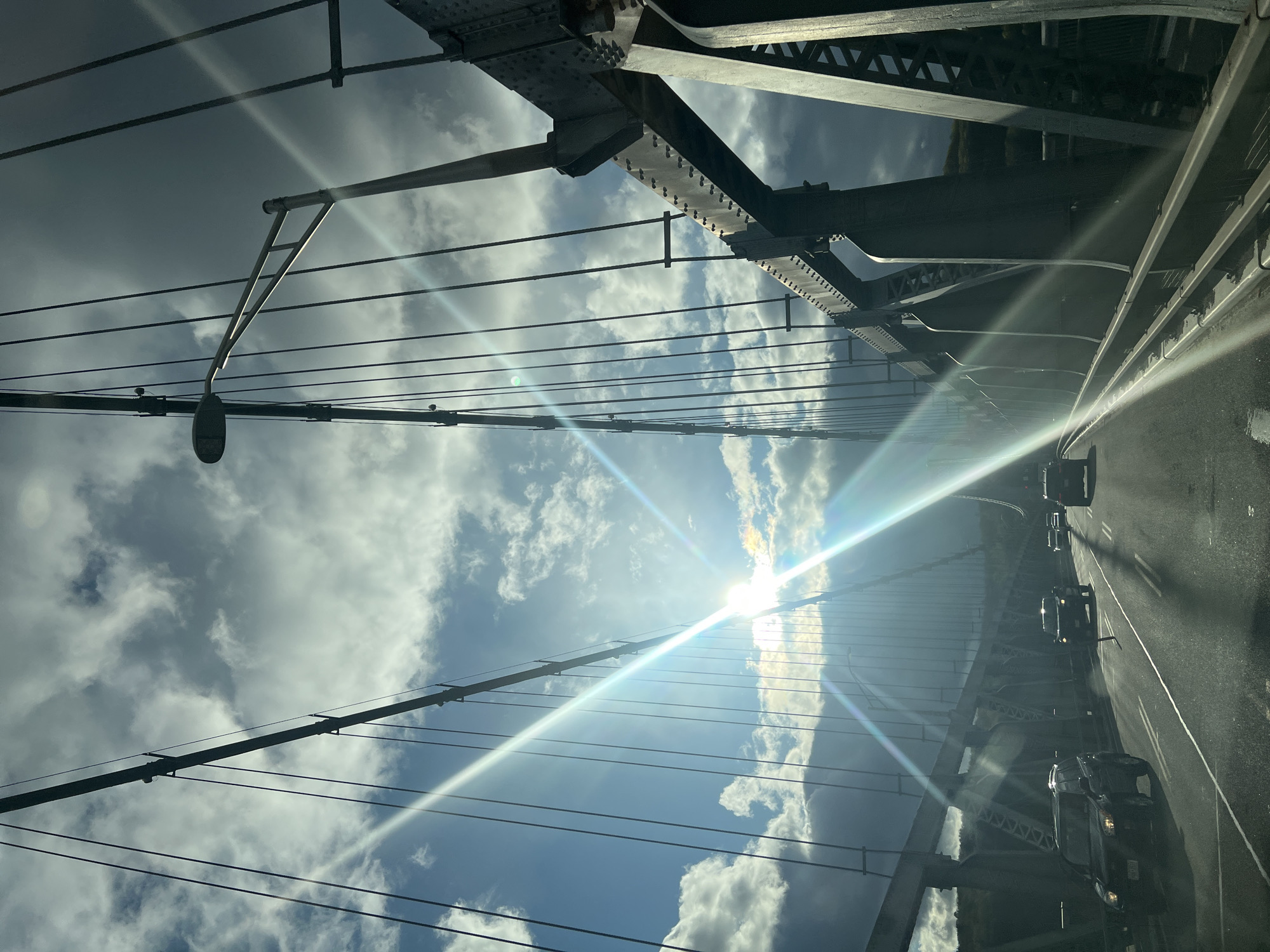 The image captures a bright, daytime scene looking forward and slightly upward from a vehicle crossing a large metal bridge. The sun is prominently visible in the upper-left quadrant of the frame, casting intense light rays and producing noticeable lens flares that extend across the sky and the bridge's structure.
The sky features a mix of bright blue patches and fluffy white cumulus clouds. The dominant subjects are the structural elements of the bridge, which include intricate riveted steel beams, heavy trusses, and multiple suspension cables or utility wires crisscrossing the upper and middle sections of the view. A streetlight fixture, attached to an overhead gantry, is visible on the left side, extending into the sky.
On the right side of the frame, a segment of the bridge's roadway is visible, featuring clear lane markings. Several vehicles, primarily dark-colored cars and SUVs, are present on the road, moving in the direction of the camera's travel. The sunlight reflects brightly off the road surface and the vehicles.
The scene depicts regular vehicular traffic on a substantial bridge under clear but partly cloudy conditions. The robust, riveted steel construction suggests an older, durable bridge design. No visible text is legible within the image, including on the partially obscured license plates of the cars. The intense sunlight indicates a sunny day, likely mid-morning or late afternoon, given the angle of the sun. The structural details align with the characteristics of a major bridge, such as the Mid-Hudson Bridge in the City of Poughkeepsie.