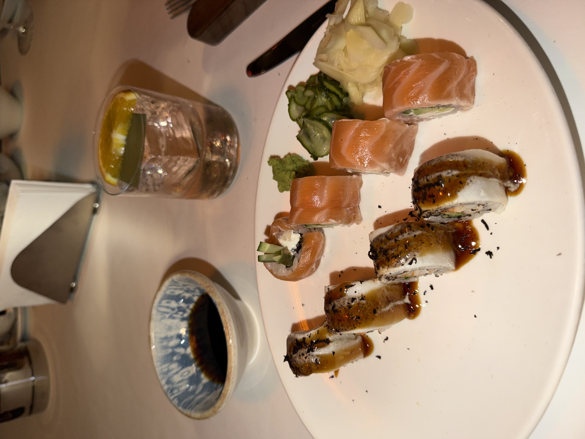 The media file displays a top-down view of a dining table in what appears to be a restaurant setting in Moscow, Russia, likely during the evening due to the ambient, warm lighting.
The central subject is a white, oval-shaped plate laden with various sushi rolls and accompanying condiments. On the plate, there are two distinct types of sushi rolls. One type consists of several pieces wrapped in thin slices of salmon, revealing fillings such as cream cheese and cucumber. The other type, arranged diagonally across the plate, features rolls topped with a white ingredient (possibly unagi or another type of fish) and generously drizzled with a dark, glossy sauce, garnished with small, dark flakes.
Adjacent to the sushi, the plate holds dollops of bright green wasabi, a serving of thinly sliced pickled ginger, and a portion of sliced pickled cucumber. To the left of the plate, a clear glass contains water with a fresh slice of orange floating inside. Below the glass, a small, ceramic bowl with a blue and white patterned interior is filled with dark soy sauce.
Other objects visible on the table include a metal napkin holder or dispenser partially obscured in the lower left corner, and cutlery (a fork and a knife) visible in the upper part of the image, suggesting a full place setting. The table surface is a smooth, light-colored material. No people are visible in the frame, and there is no discernible text. The scene captures a prepared meal in an indoor, intimate dining environment.