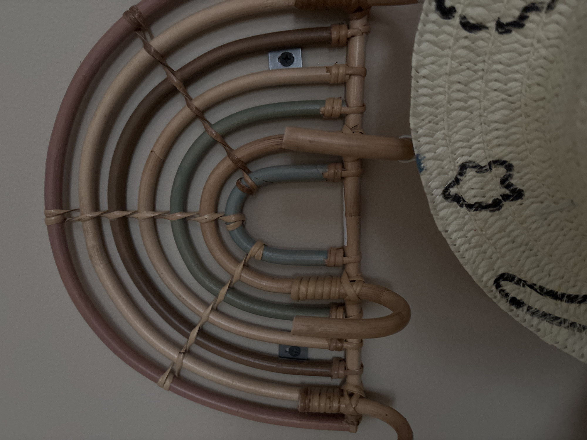 A close-up, eye-level, indoor shot features a rustic, rainbow-shaped wall rack made of rattan. The rack has multiple arched segments in muted colors, including pink, beige, brown, and light green, with some segments painted a soft blue. The rack is secured to a plain, light-colored wall with two visible screws. Several wooden hooks protrude from the bottom of the rack, designed for hanging items. Below the rack, a portion of a woven straw hat is visible, decorated with simple black star-shaped patterns. The lighting is soft and even, suggesting an indoor environment. No specific geographical markers are present to identify a location in Dubai, United Arab Emirates.