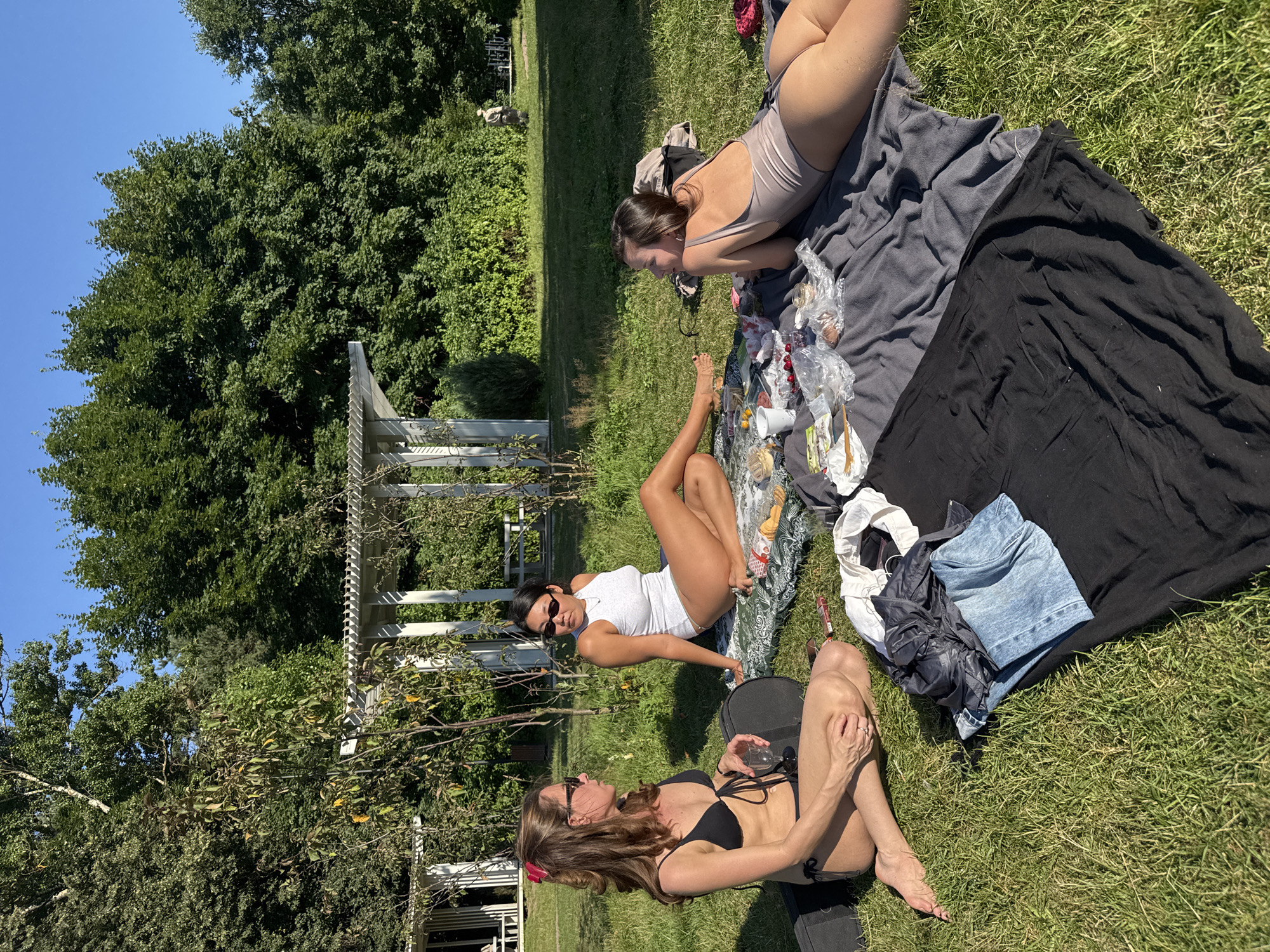 Three women are having a picnic on a grassy area on a sunny day. One woman in a black bikini is sitting on the left, holding a glass and looking at another woman. The second woman is wearing a white tank top and sitting on a picnic blanket in the center. The third woman is lying on her stomach on a gray blanket on the right, wearing a taupe swimsuit. The picnic blanket has food and drinks spread out on it, including plastic containers of food, a plastic cup, and what appears to be fruit. Clothing is scattered on the grass near the blankets. In the background, there is a white gazebo-like structure, trees, and some small buildings. The scene appears to be set in a park or recreational area. There is no indication that this location is in Moscow, Russia.
