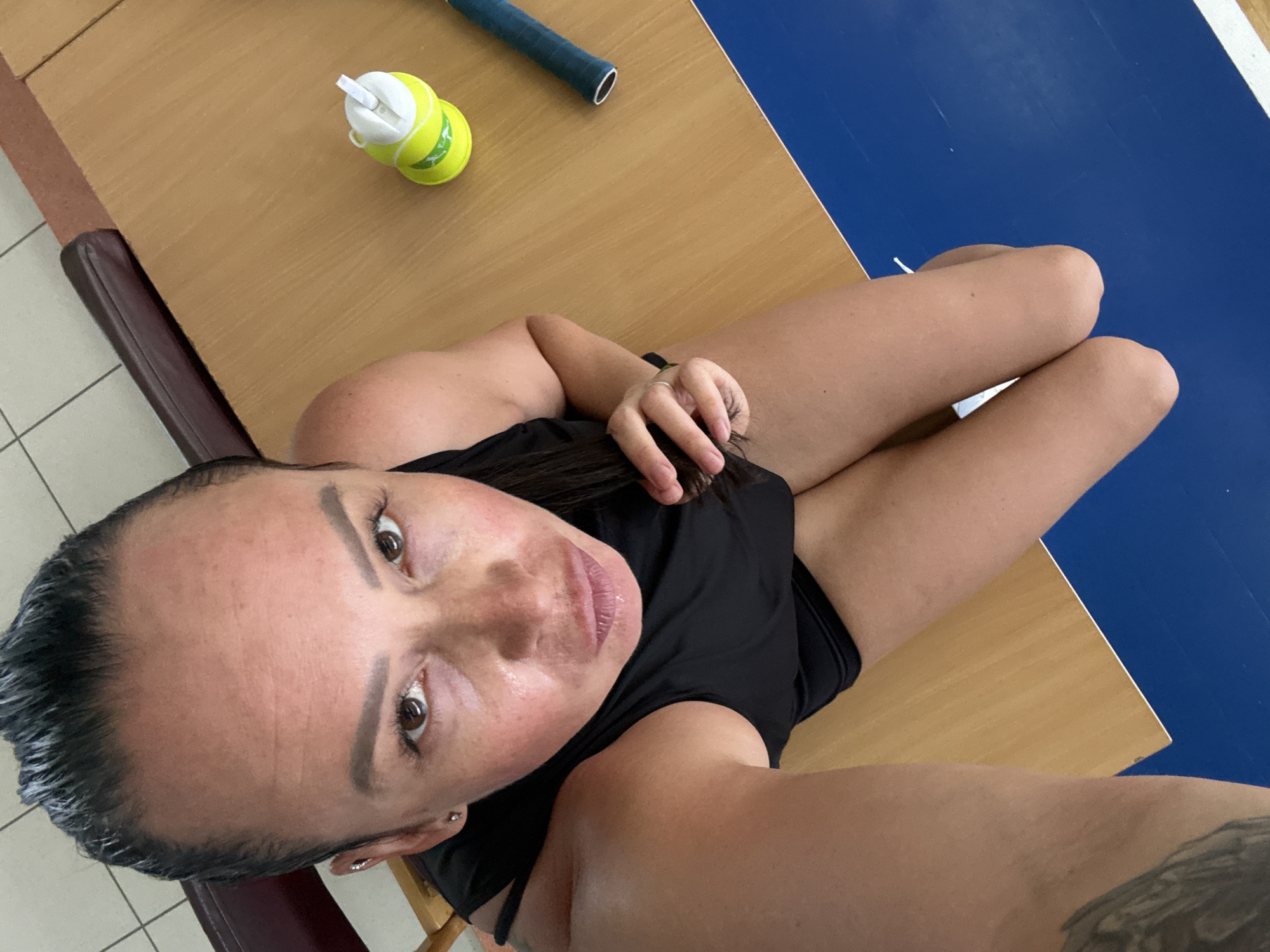 In a top-down shot, a woman with dark hair pulled back sits on the floor, her knees bent and legs crossed. She wears a black tank top and dark shorts. Her gaze is directed upwards at the camera, and her lips are slightly parted. A water bottle and a tennis racket lie on a wooden surface next to her. The background suggests an indoor sports facility, possibly a gymnasium, with a blue mat on the floor and a brown bench. The overall scene implies a break during a sporting activity. The presence of a tennis racket indicates a possible location such as a tennis court or a sports hall in Chaikovsky, Russia.