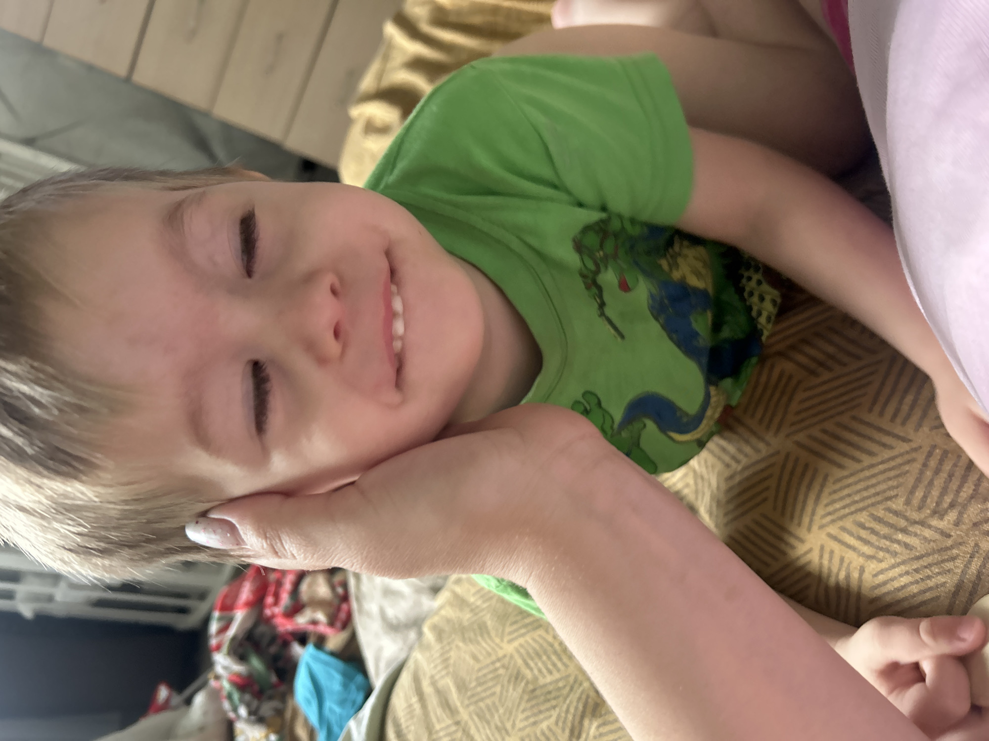 A young child, likely a toddler, is lying down on a bed or sofa, wearing a green T-shirt with a blue and green design. The child's eyes are closed and they are smiling slightly. A hand is supporting their head. The bedding is patterned in yellow and brown. In the background, partially obscured, are what appear to be a radiator and other household items. It is impossible to verify the location as Chaikovsky, Russia from this image.