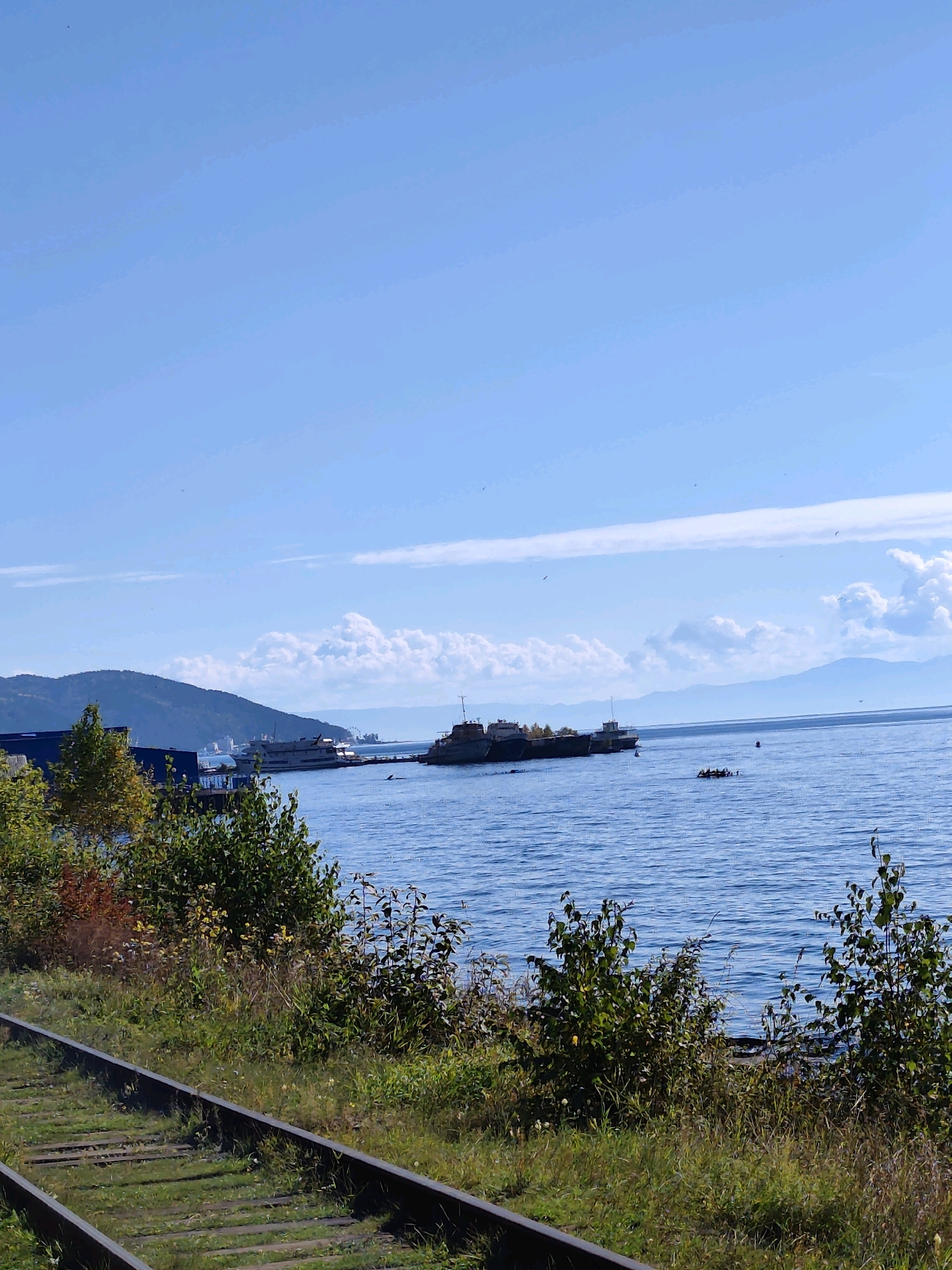 The image shows a scenic view of a body of water, likely a lake, with several boats docked along the shore. In the foreground, a set of train tracks runs parallel to the water's edge, with lush green grass and trees growing alongside. The water is calm and blue, reflecting the clear sky above. Distant mountains form a picturesque backdrop to the scene. A few fluffy white clouds are scattered across the sky. It appears to be a sunny day with good visibility. The overall impression is one of natural beauty and tranquility, with a hint of industrial presence from the docked boats and train tracks.